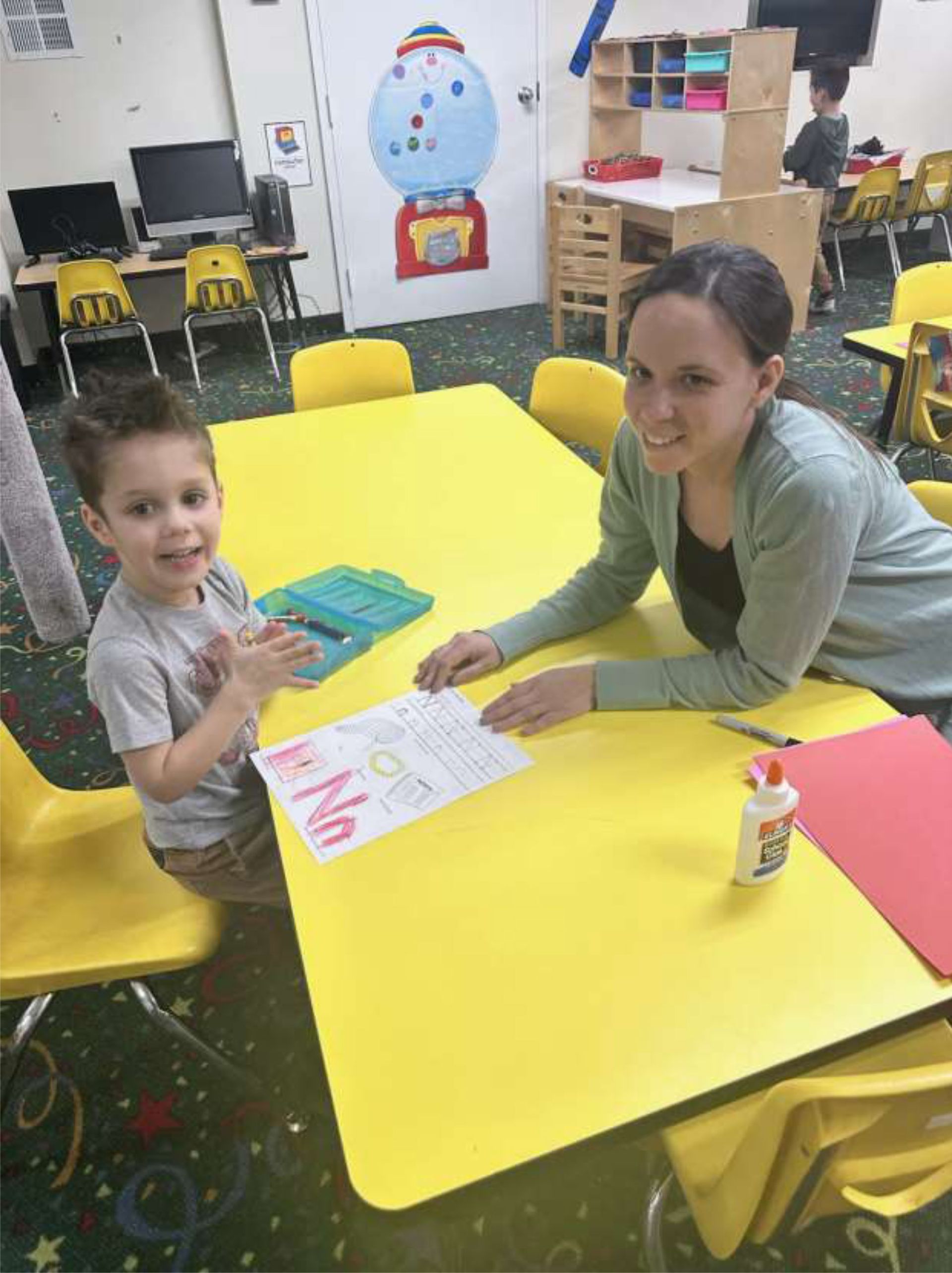 A young boy and woman sit at a yellow table in a classroom, working on an art project with the letter 