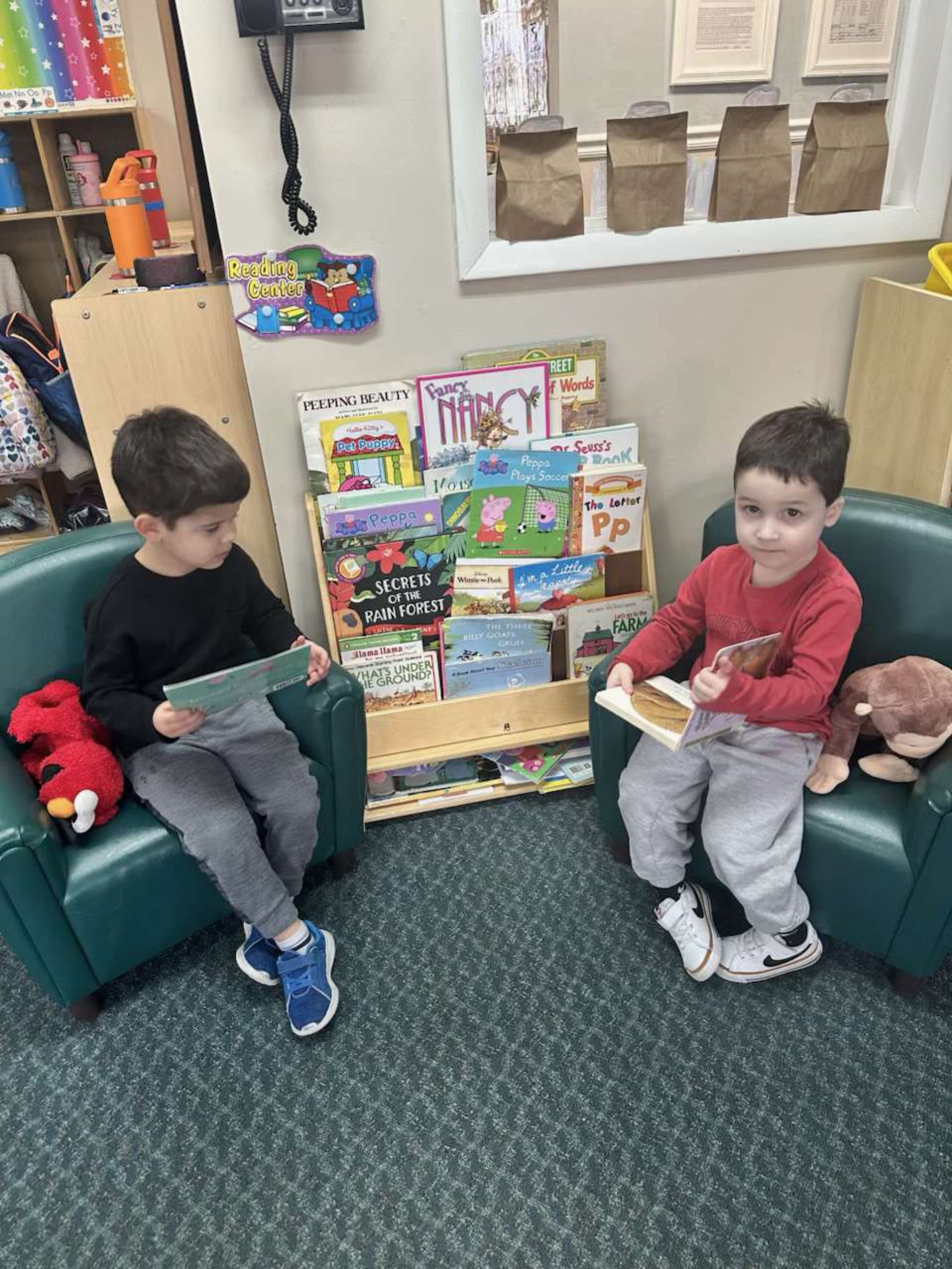 Two young boys in casual clothes sit in green armchairs, reading books in a brightly lit room with a bookshelf.