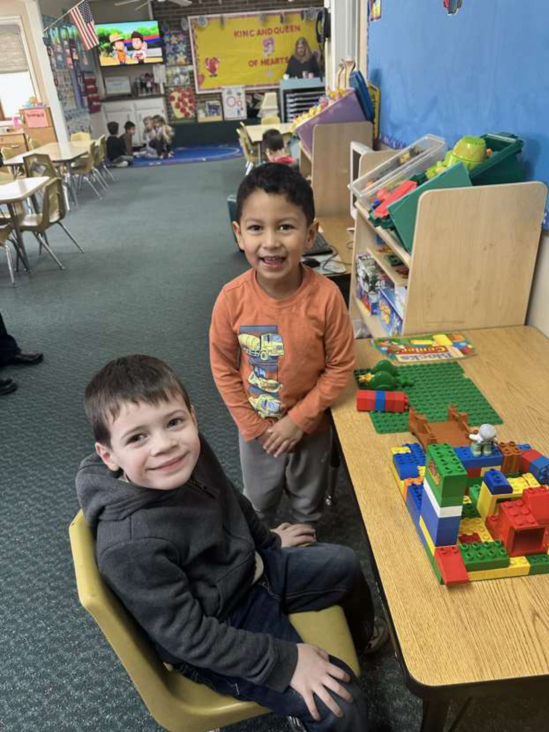 Two young boys smile at the camera. One sits in a chair, the other stands beside a table with a LEGO structure on it, in a classroom setting.