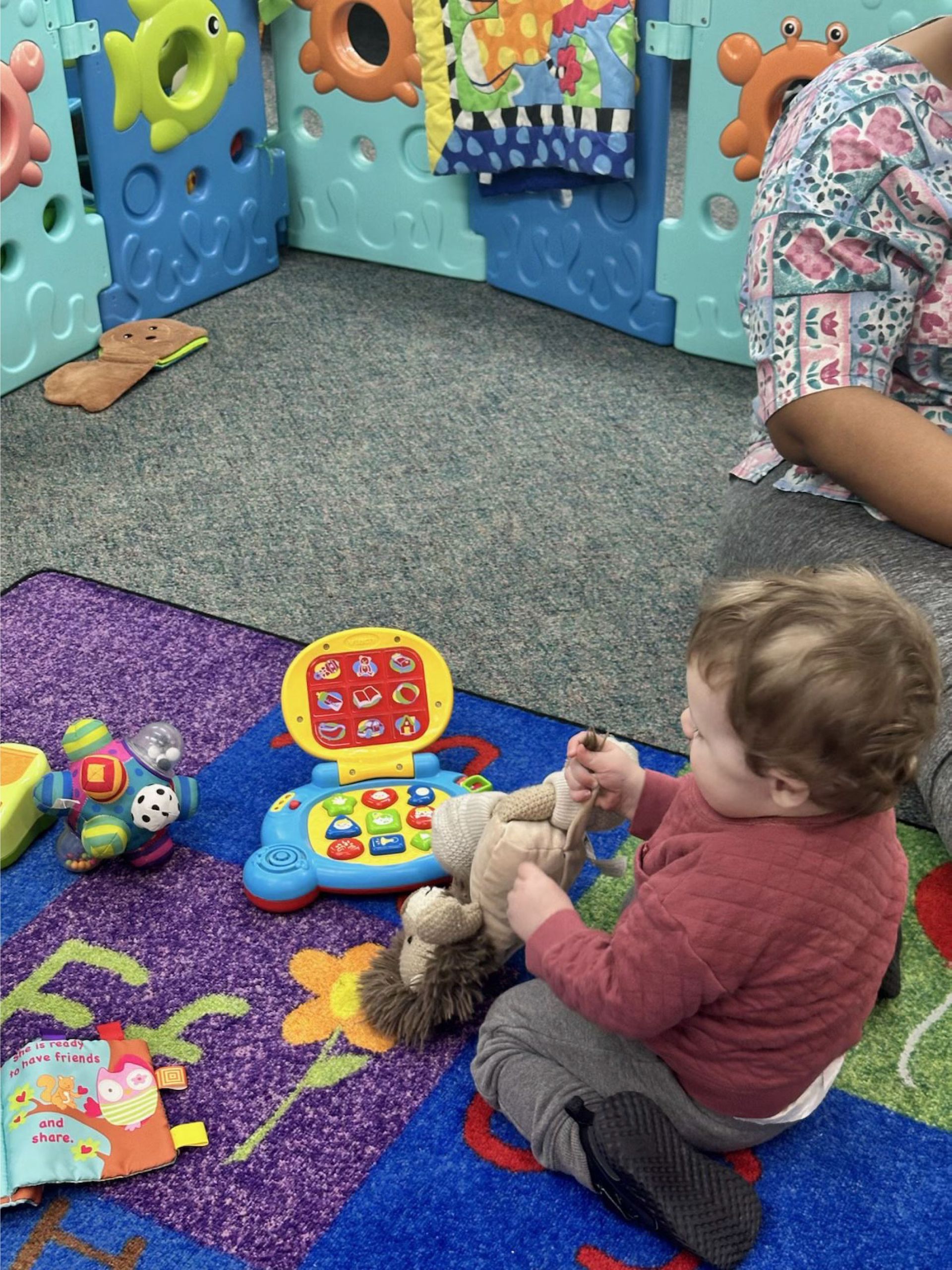 A young child in a red shirt plays with toys on a colorful rug, including a stuffed lion and a toy laptop, in a play area