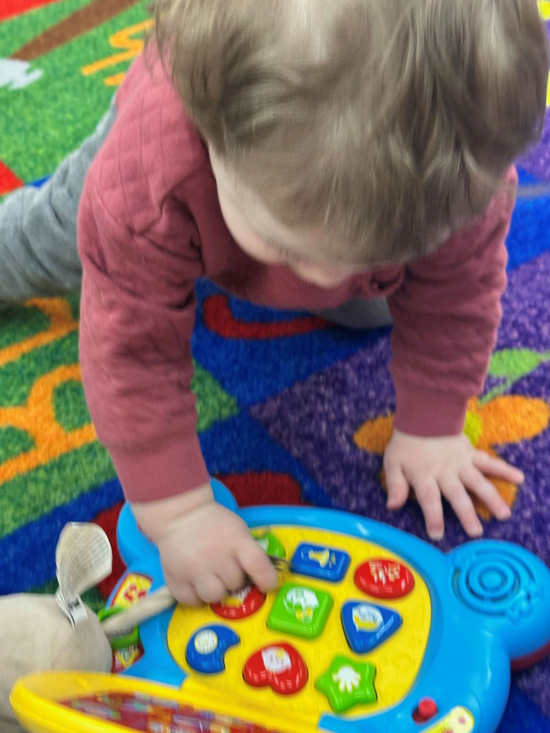 A toddler plays with a colorful toy on a patterned rug, reaching with one hand to press the shapes