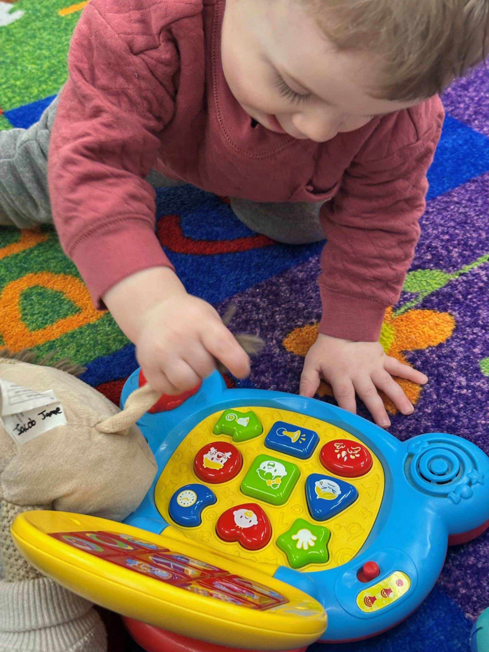 Toddler playing with a colorful, shaped button toy on a patterned carpet, reaching with a finger