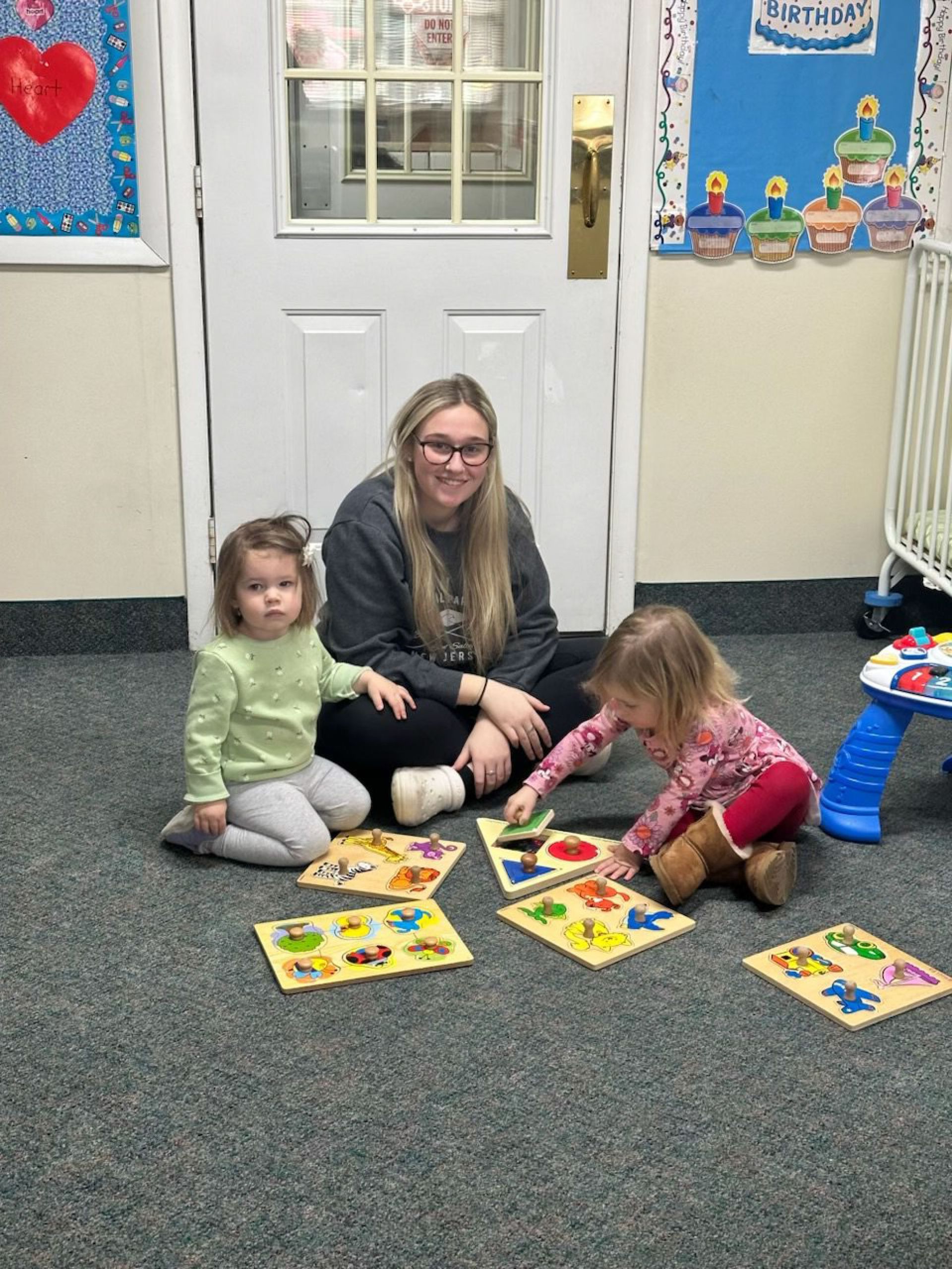 A woman and two toddlers sit on a carpet, playing with puzzles