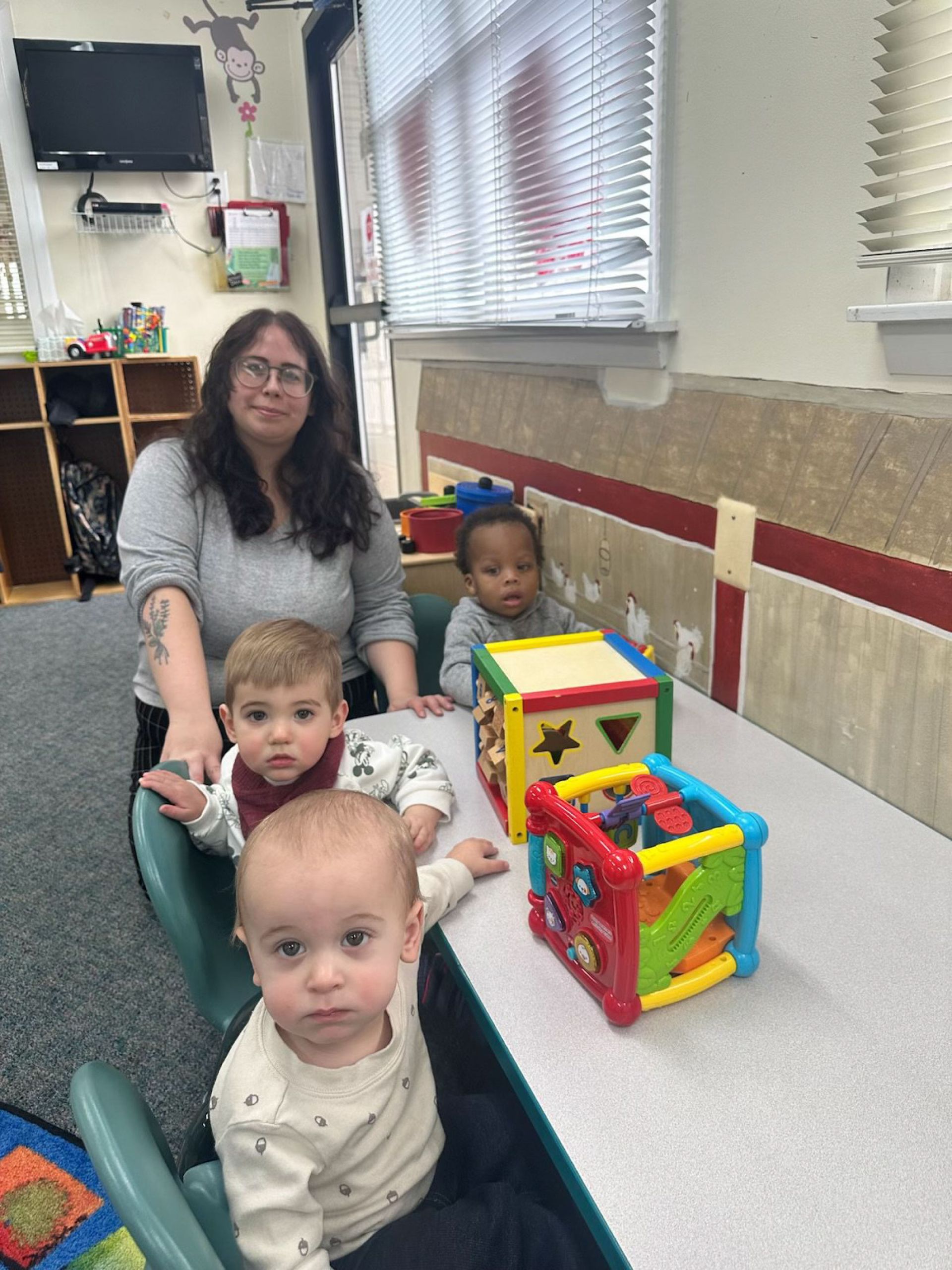 A teacher and three babies sit at a table with colorful blocks in a daycare