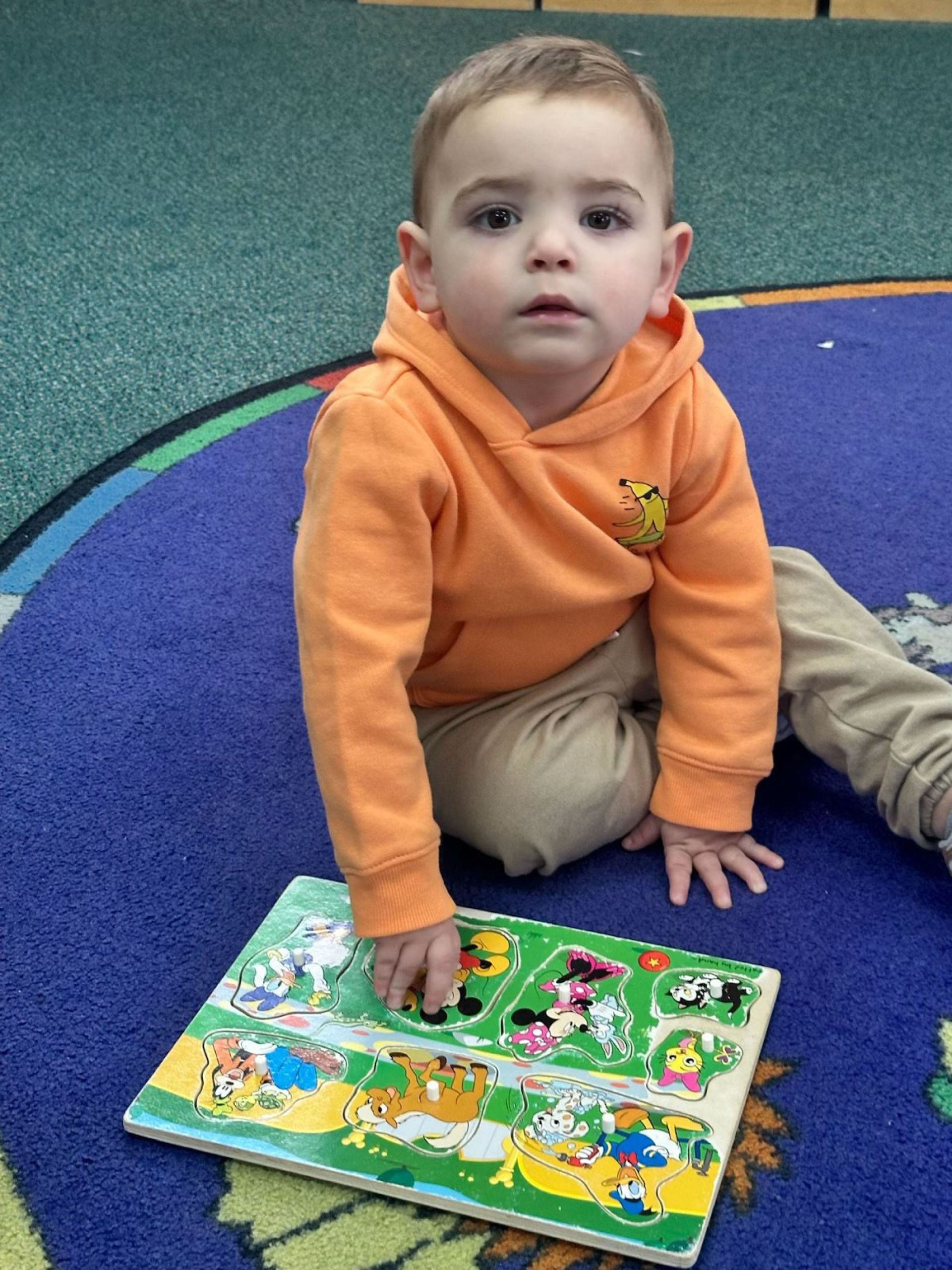 Young child in an orange hoodie playing with a puzzle on a blue carpet