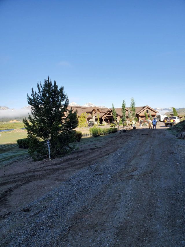 Gravel driveway leading to a large brown building with landscaping. Two people stand near the entrance; mountains in the distance.