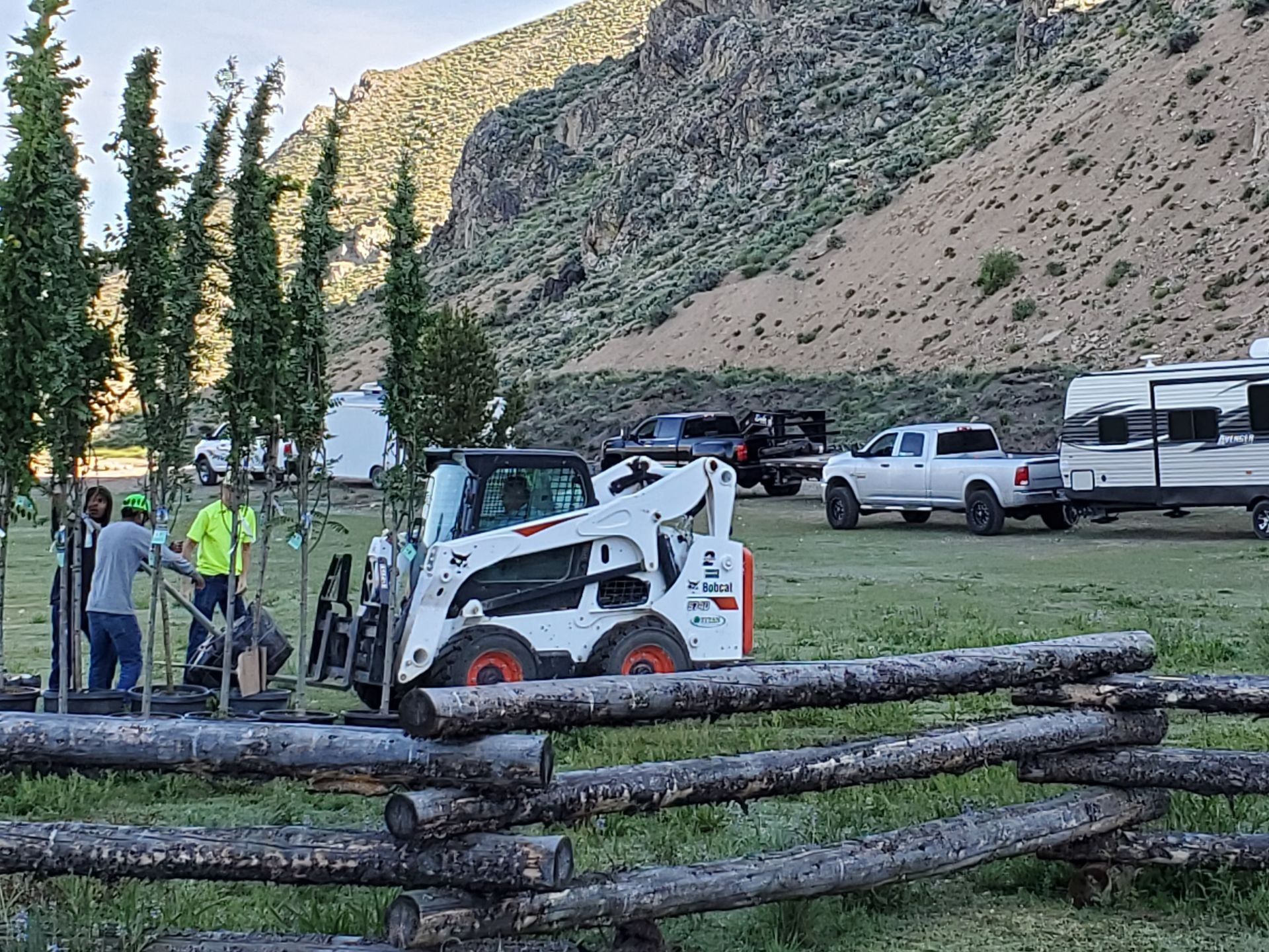 Bobcat working near fence; workers planting trees. Trailers and vehicles parked, mountains in background.