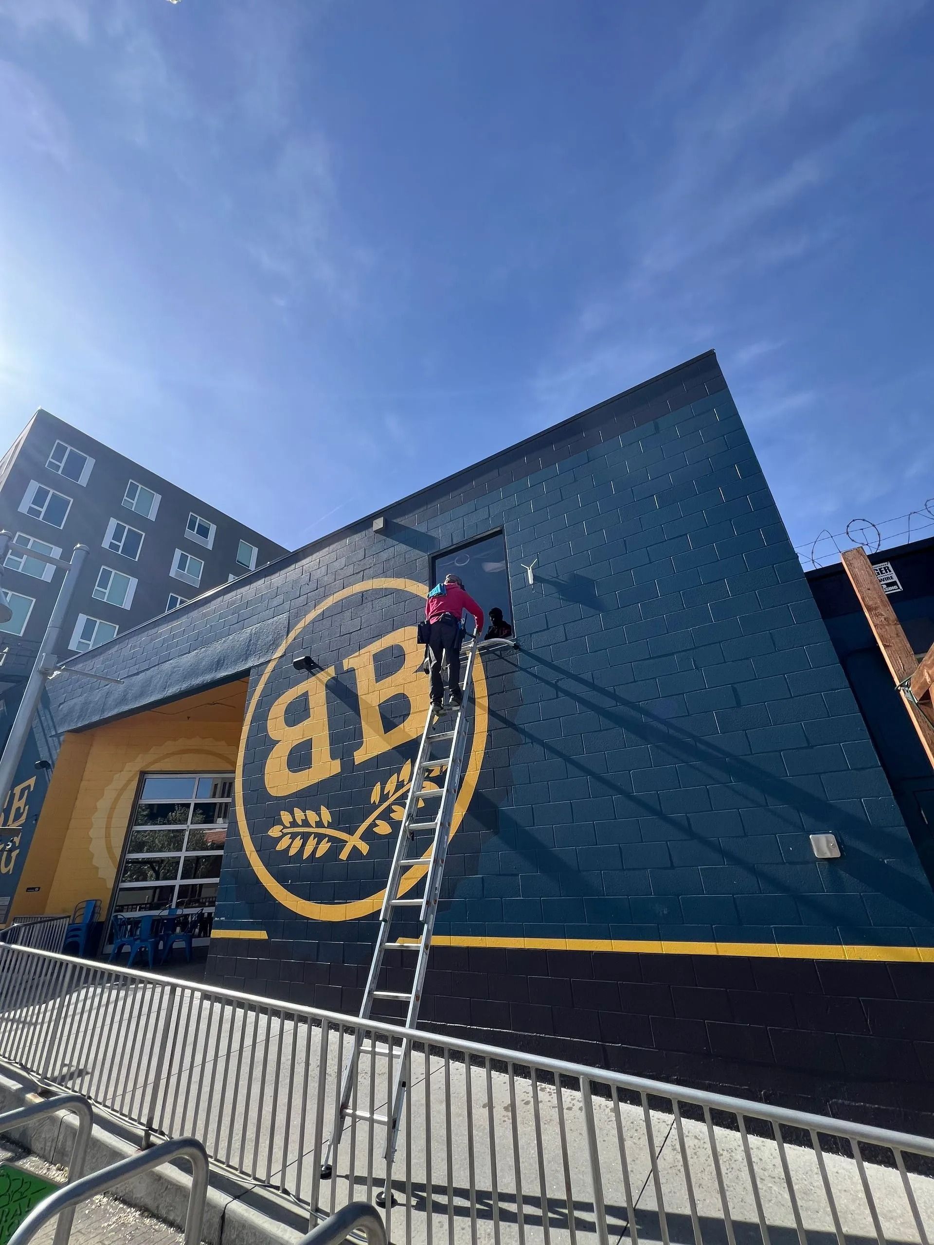 Person on a ladder, working on a dark blue building with a large logo, under a clear sky.
