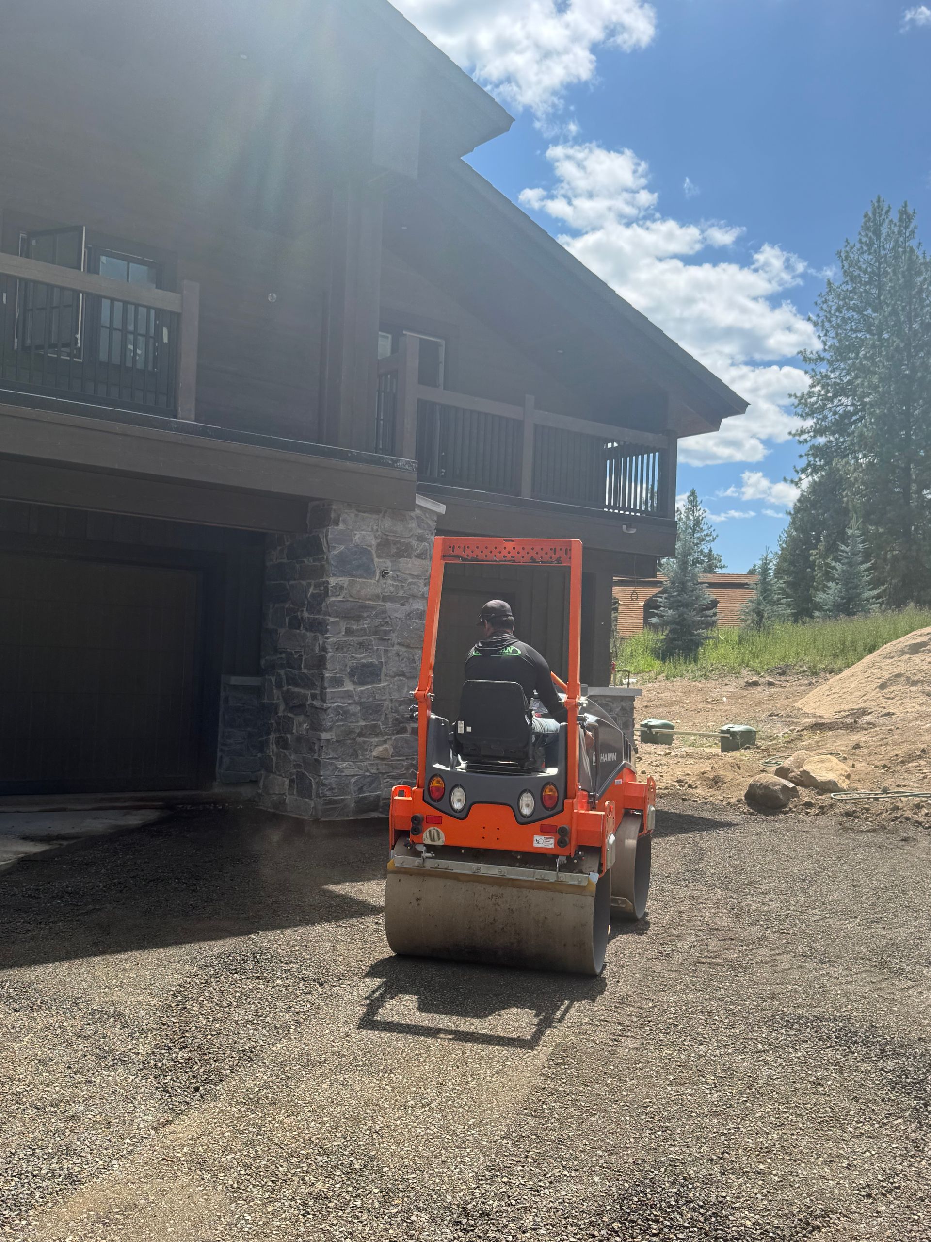 Orange roller compactor on gravel driveway in front of a wood-sided house; operator visible.