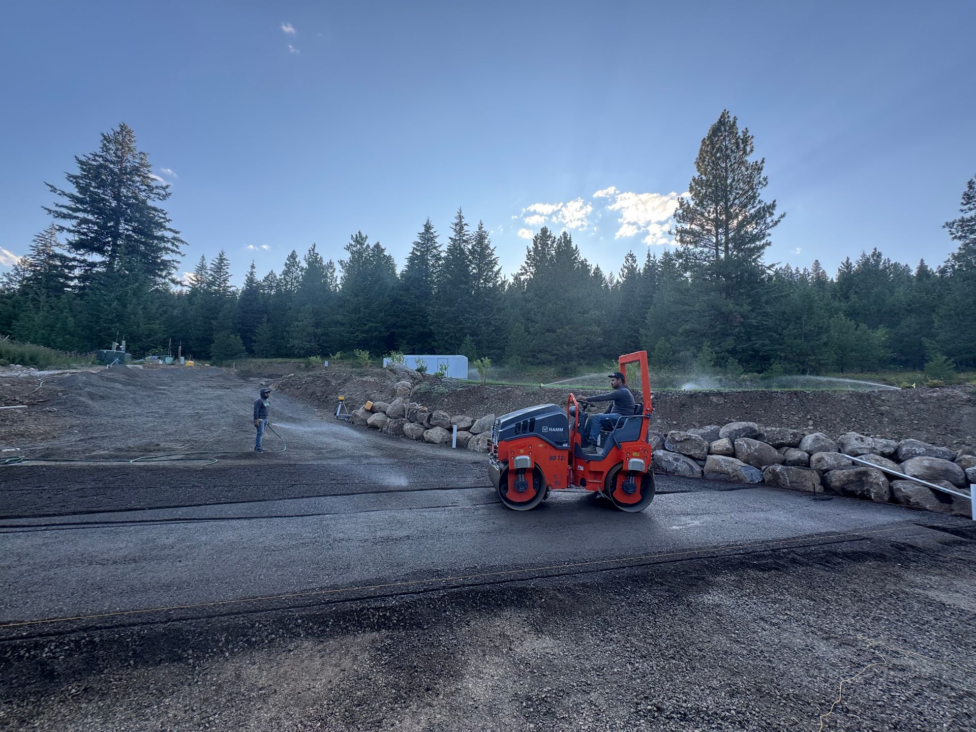 A construction worker operating a compact roller on a gravel surface under a blue sky, surrounded by trees.