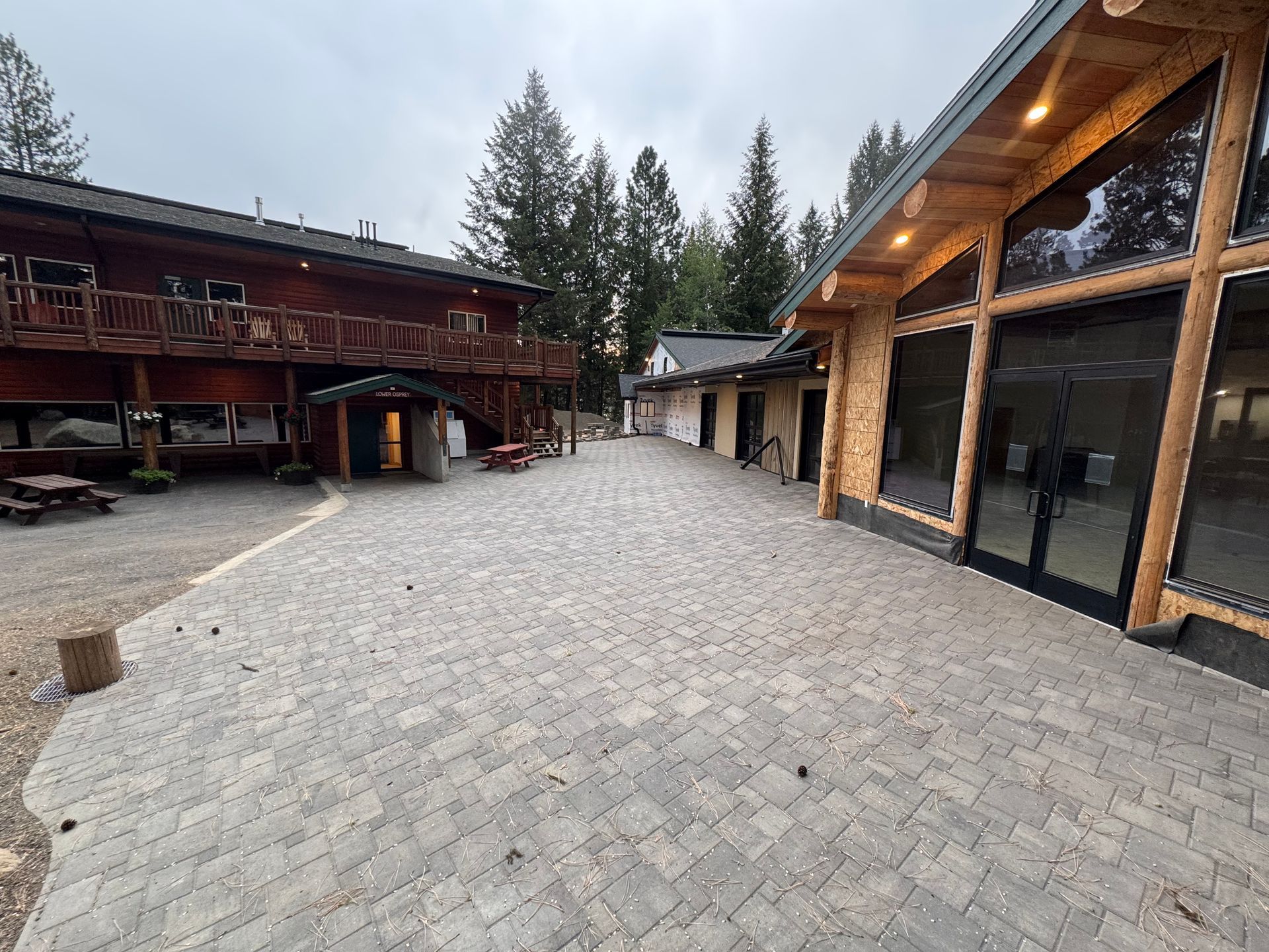 Outdoor courtyard with brick pavers, flanked by wooden buildings and trees. Overcast sky.