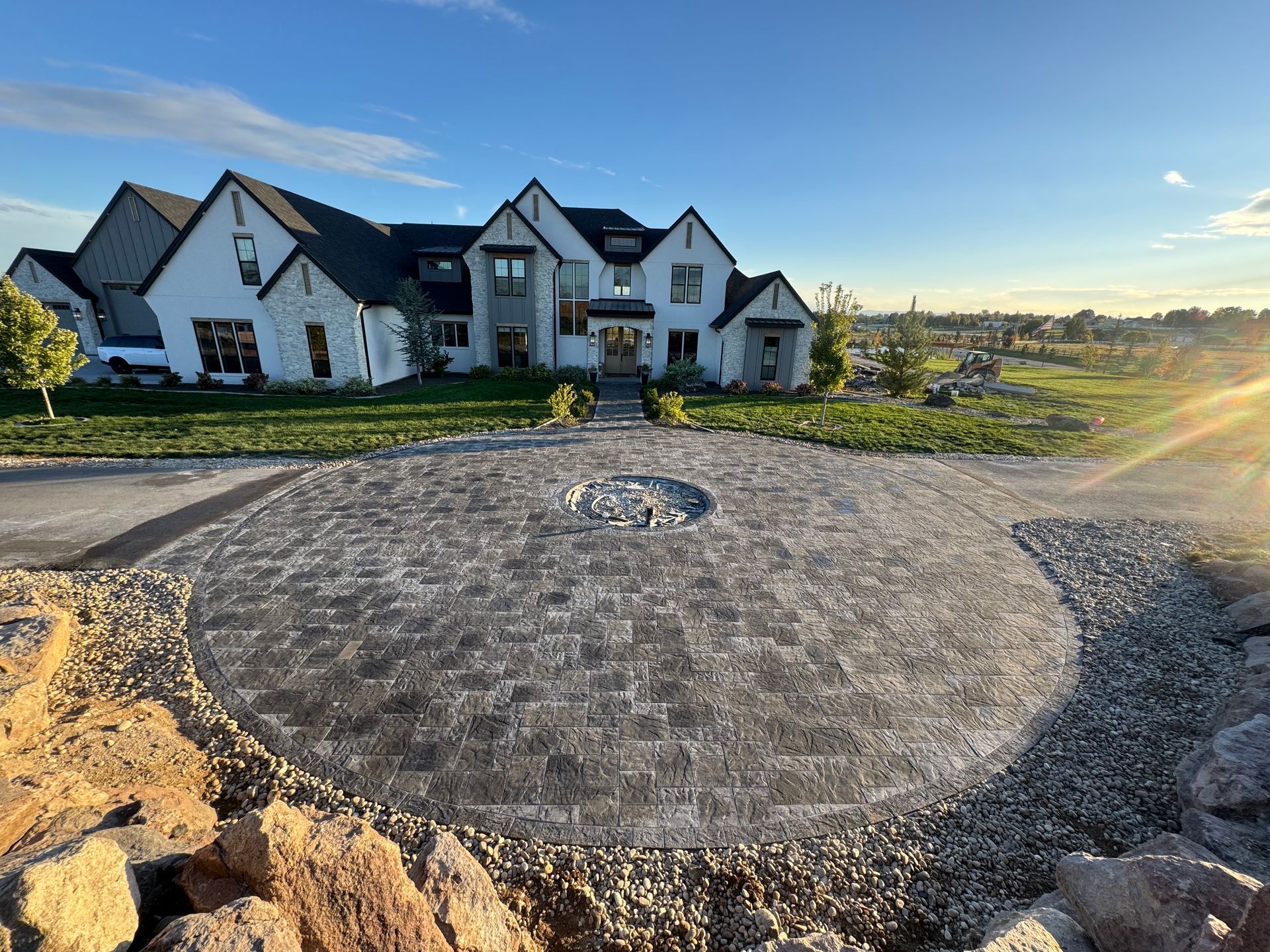 Large house with circular paver driveway, gravel border, and rock edging under a blue sky.
