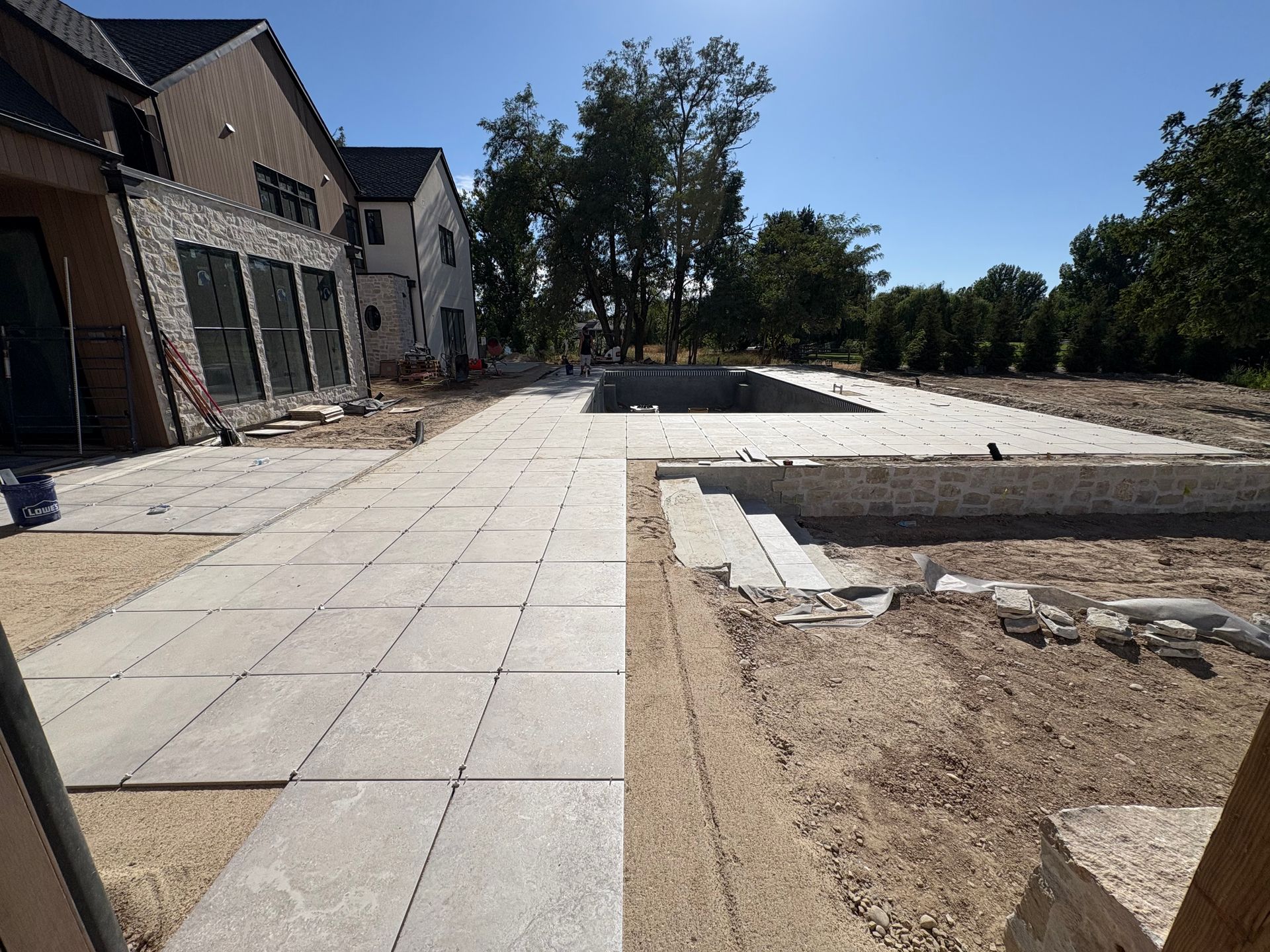 Construction site with stone patio, pool foundation, and a large house under a clear sky.