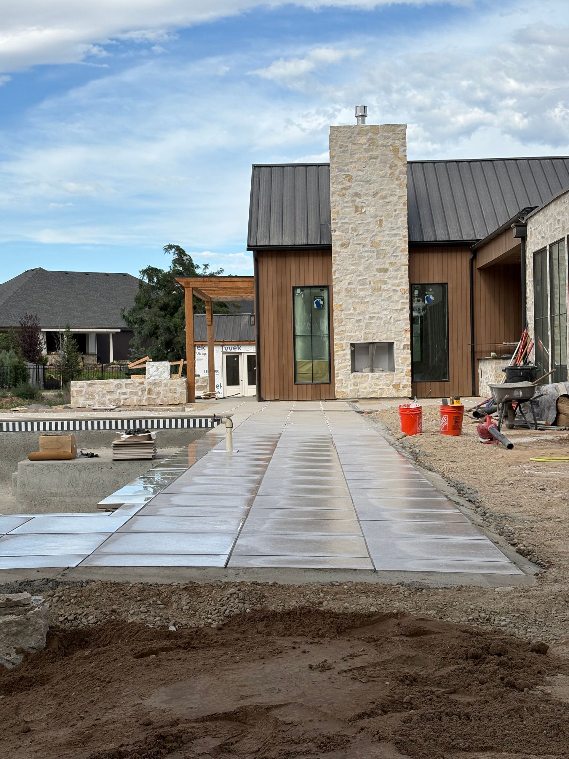 Patio construction near a house with a stone chimney and pool. Gray pavers and dirt visible.