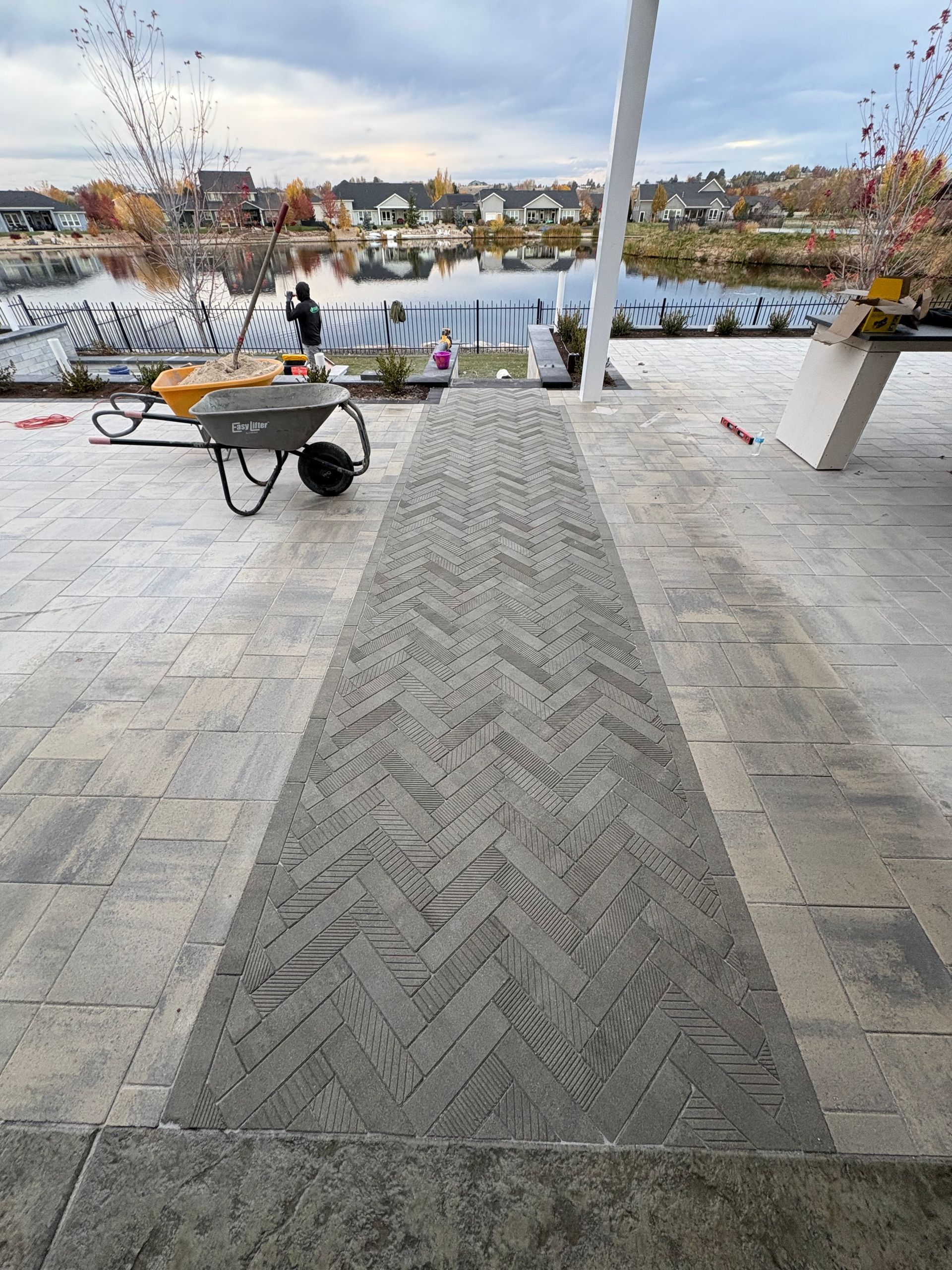 Exterior patio with patterned walkway, wheelbarrow, and waterfront view under cloudy sky.