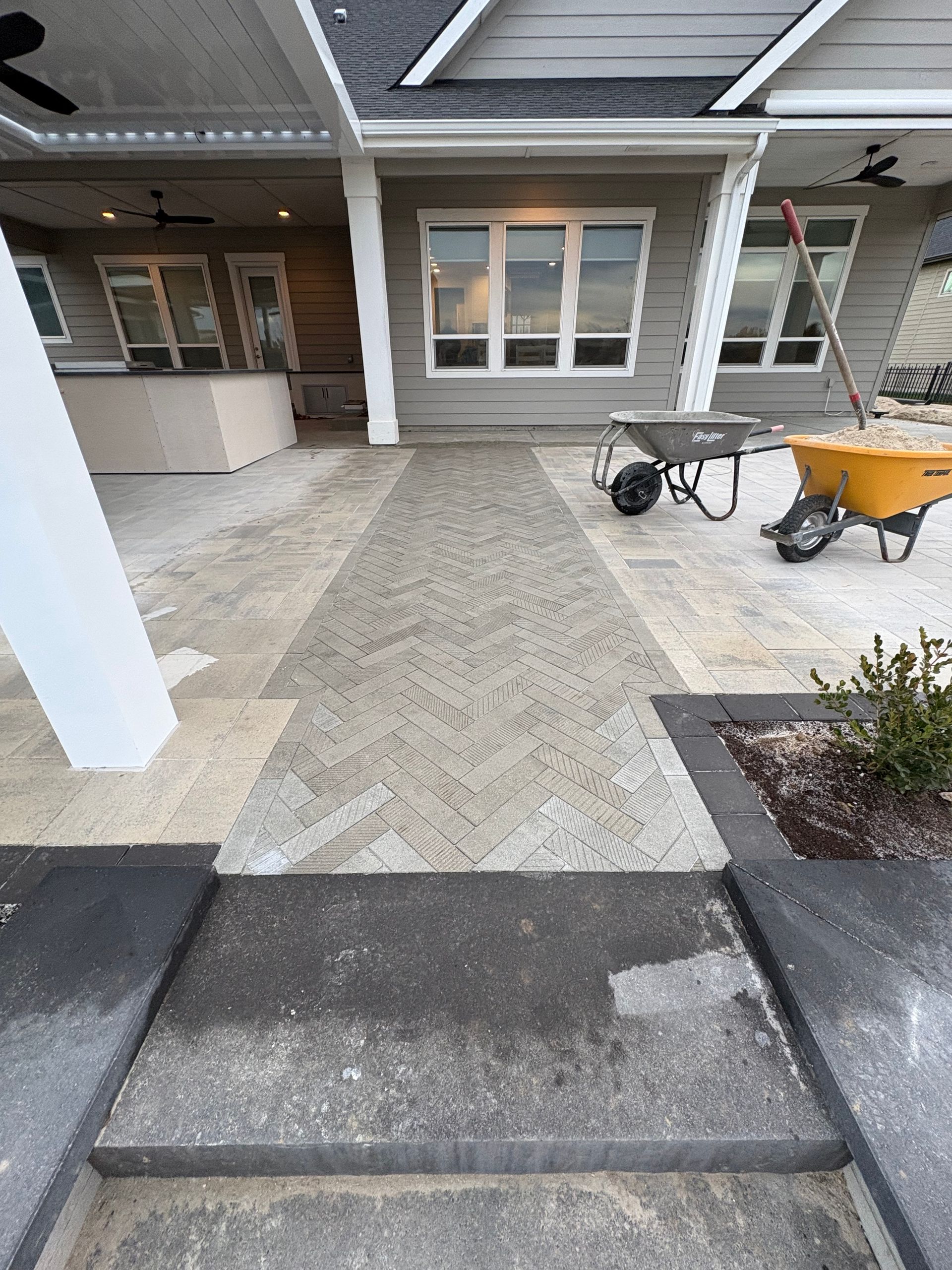 Stone patio with walkway, bordered by landscaping and the side of a house. Wheelbarrows sit nearby.