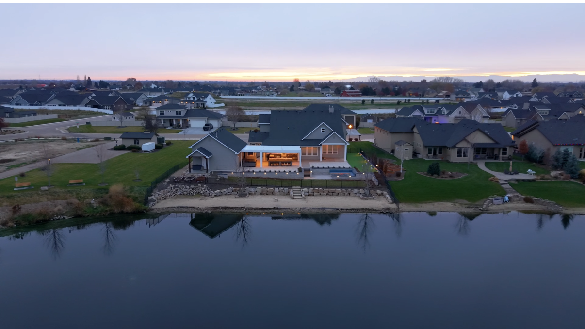 Aerial view of a house on a lake at dusk, with lights on, surrounded by other houses.
