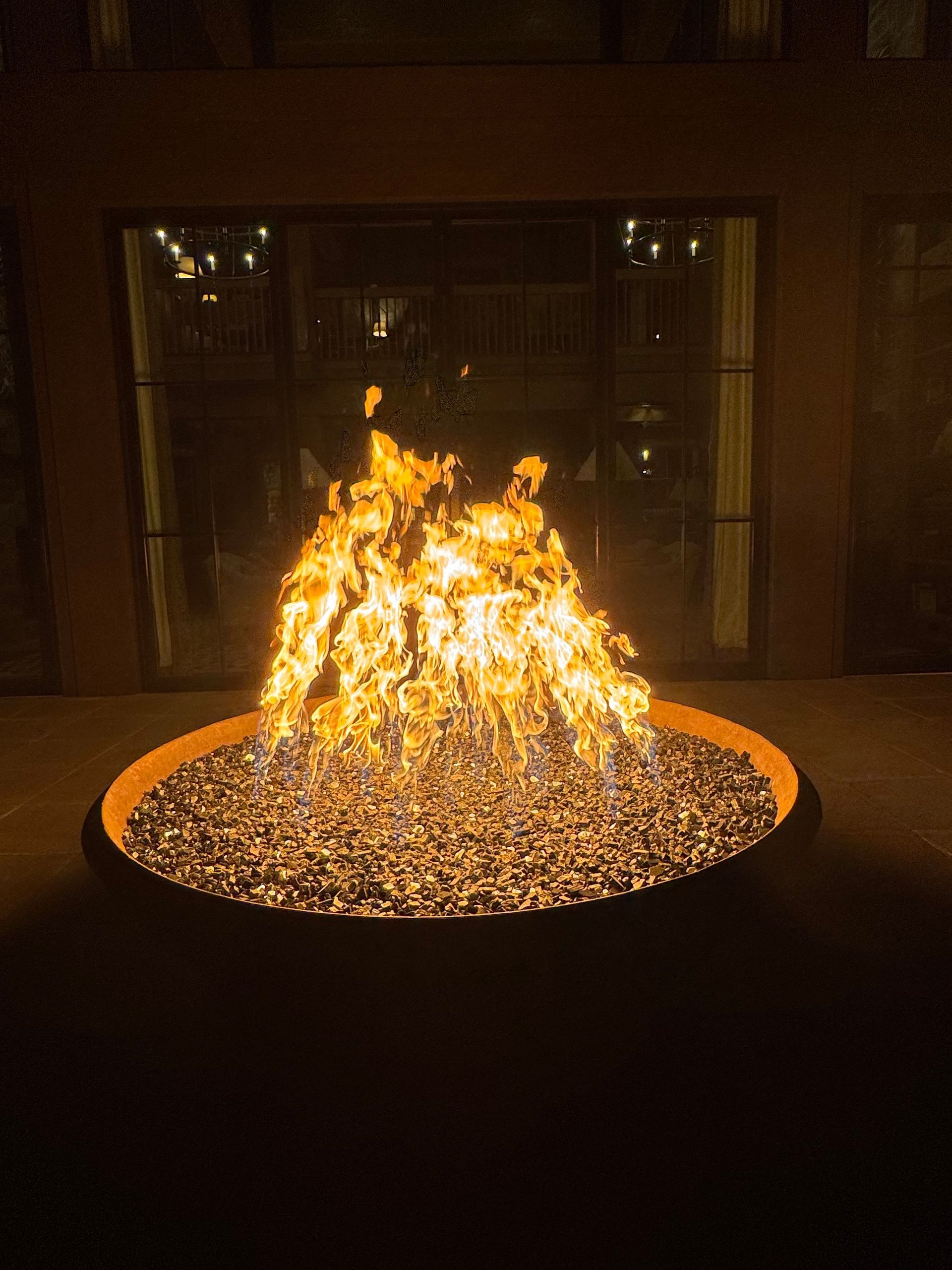 Fire pit ablaze with flames, surrounded by glowing embers, outside a building with lit windows at night.
