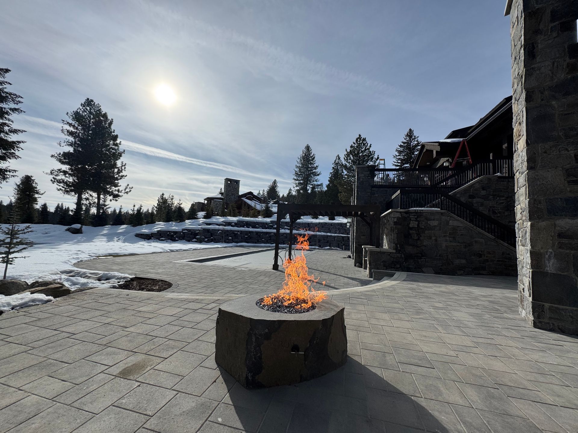Stone fire pit with flames on a paved patio, snow-covered ground, building in background, sunny sky.