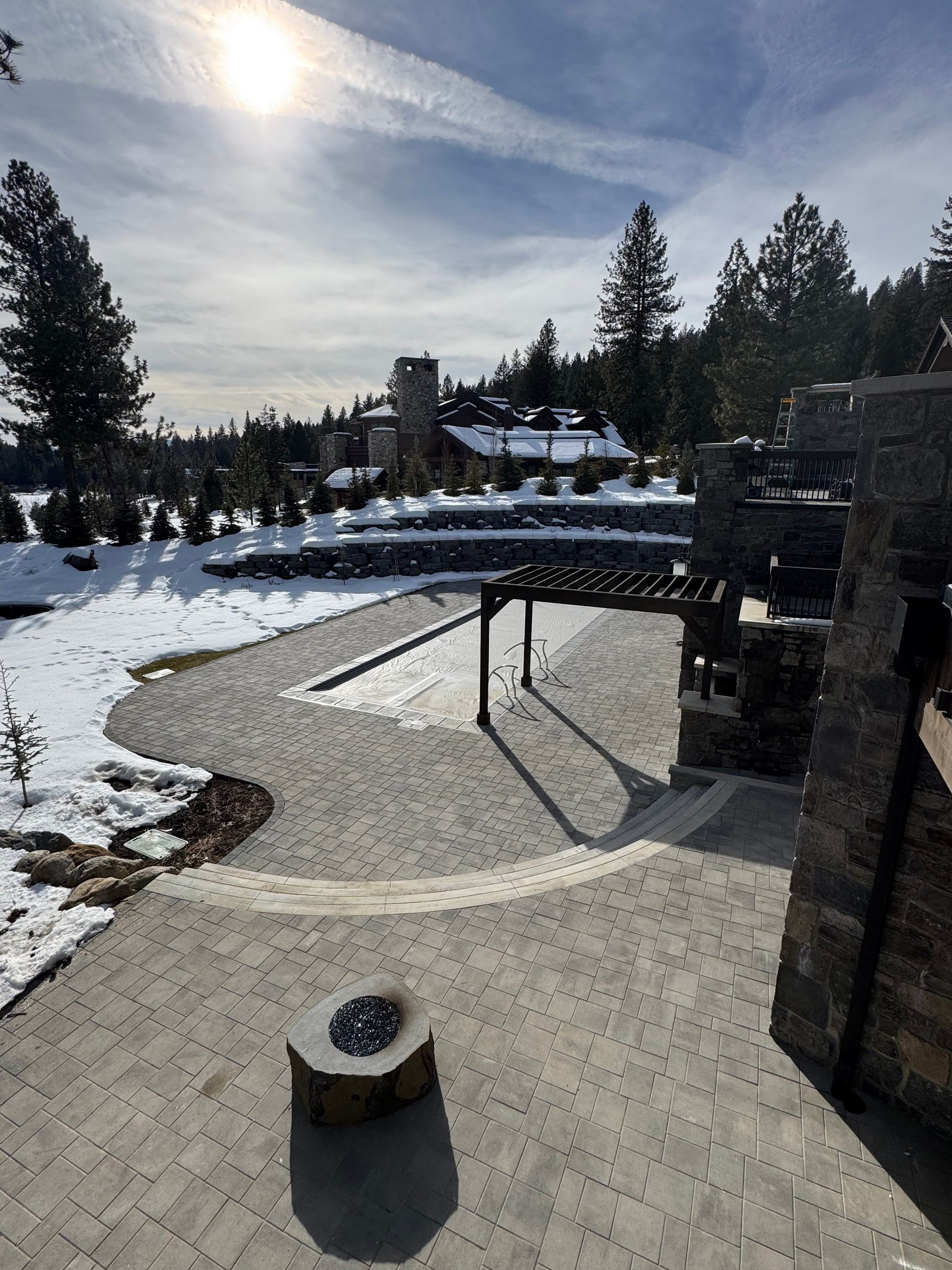 Brick patio with fire pit, stone structure, snow-covered path leading to buildings under a bright sun.