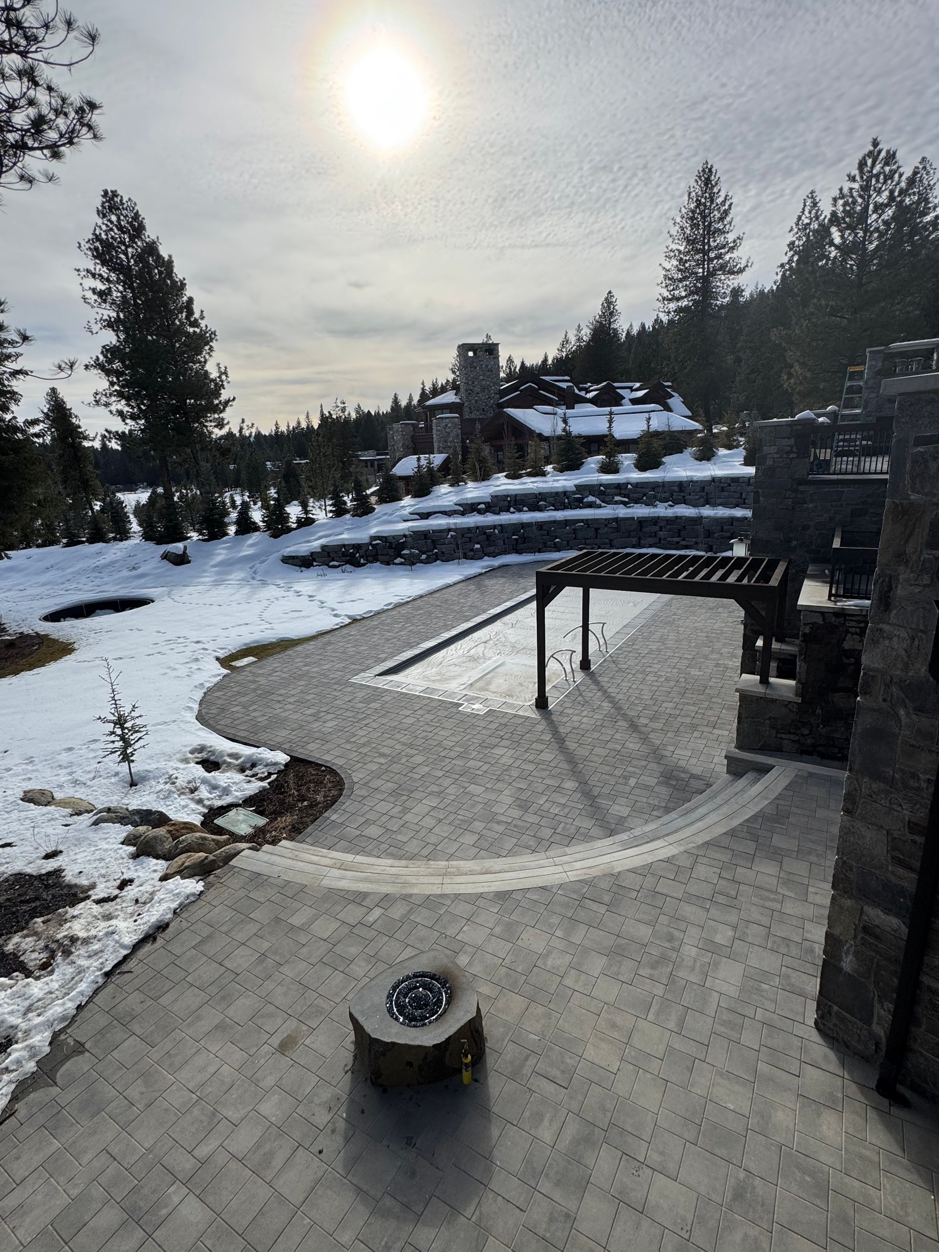 Snowy patio with paved path, fire pit, pergola, and view of a distant house with trees, under a bright sun.