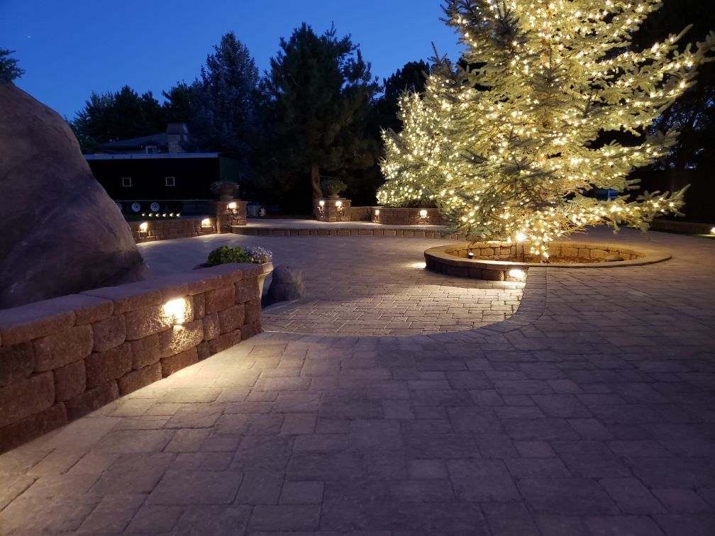 Lit brick patio with up lights and a tree decorated with string lights at dusk.