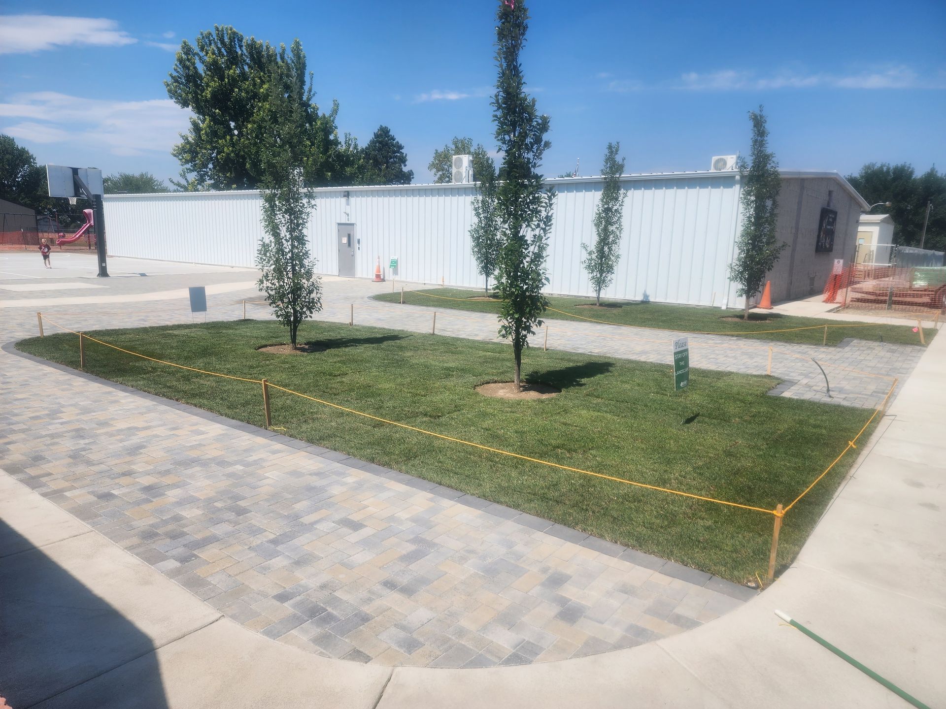 Grassy area with trees in front of a white building, surrounded by paved walkway and construction fencing.