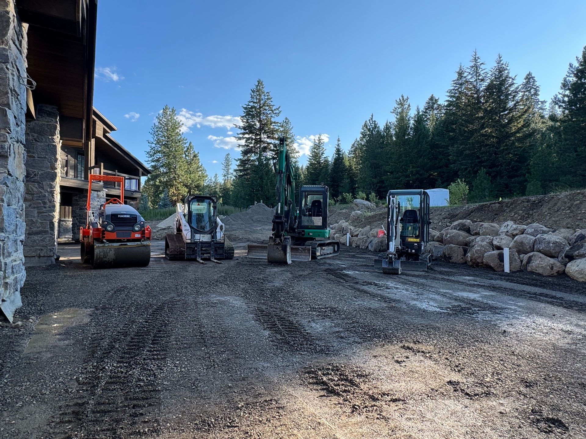 Construction site with several pieces of heavy machinery, including a roller and excavators, on a gravel surface near a building.