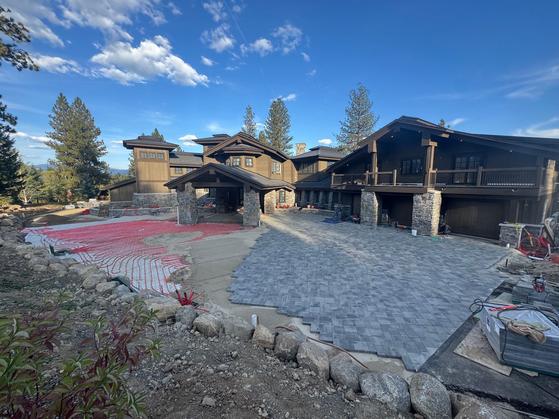 Large, rustic house under construction; driveway paved with dark stones, red material visible.