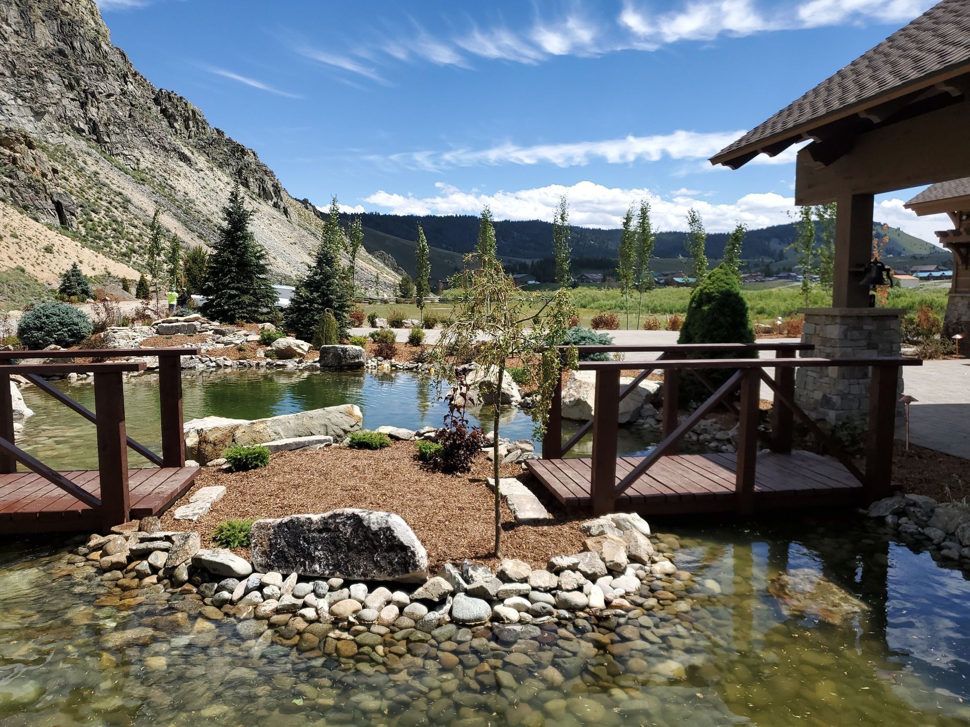Pond with wooden bridges, landscaping, and mountain backdrop under a blue sky.