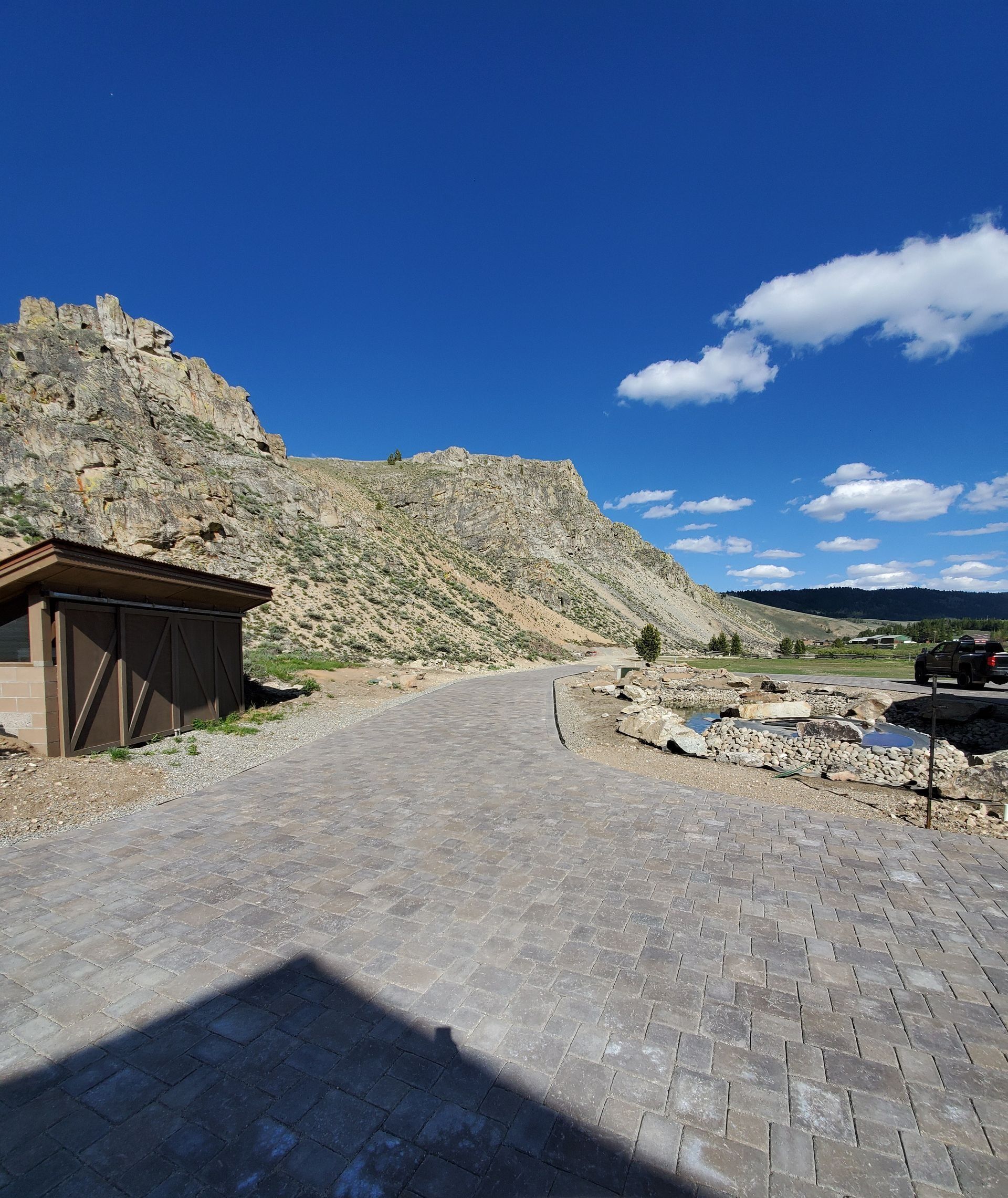 Stone path leading to a rocky mountain under a bright blue sky with sparse clouds. A small wooden structure sits on the left.