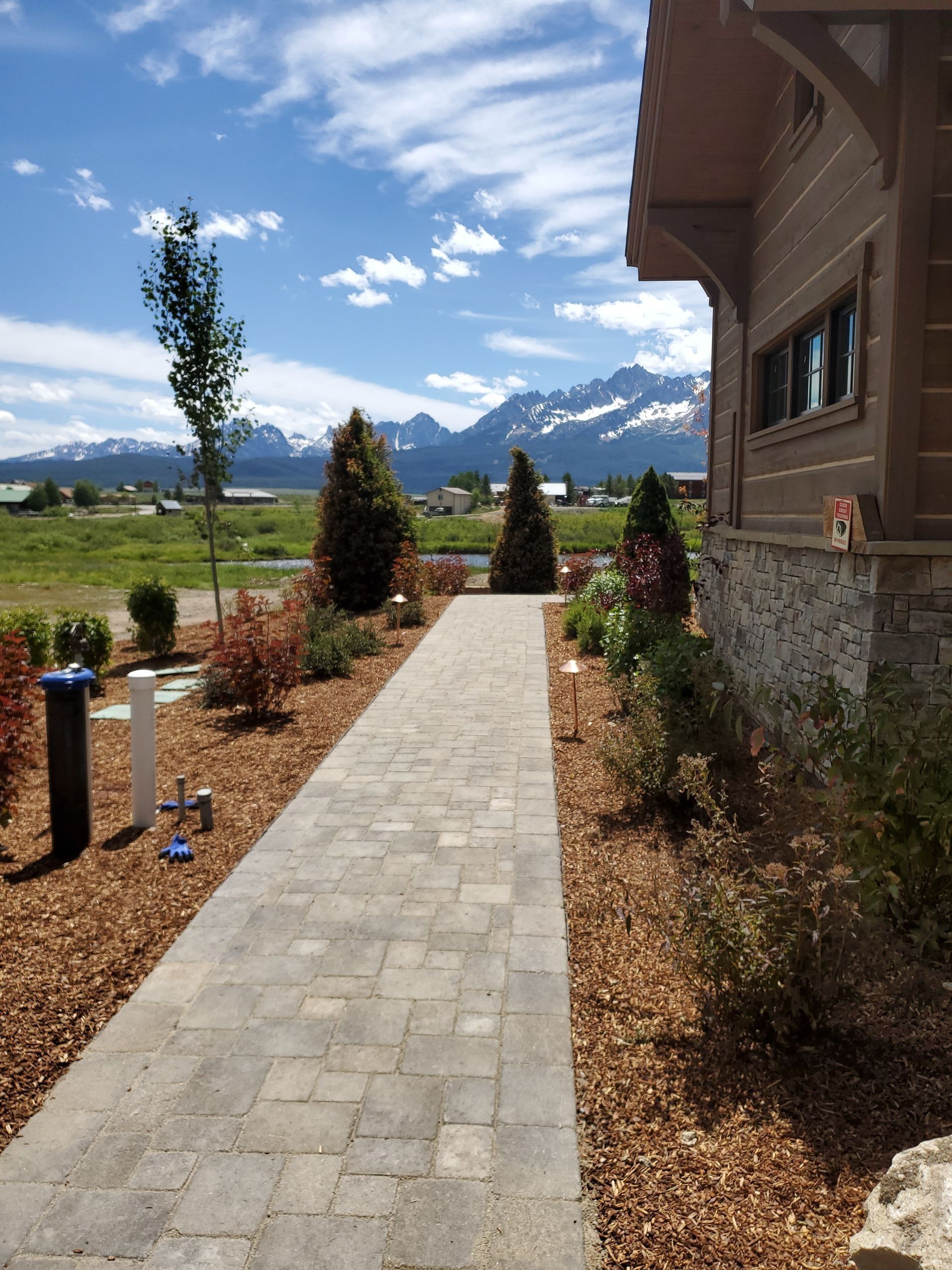 Brick pathway leading to a log cabin, with mountains in the background under a blue sky.