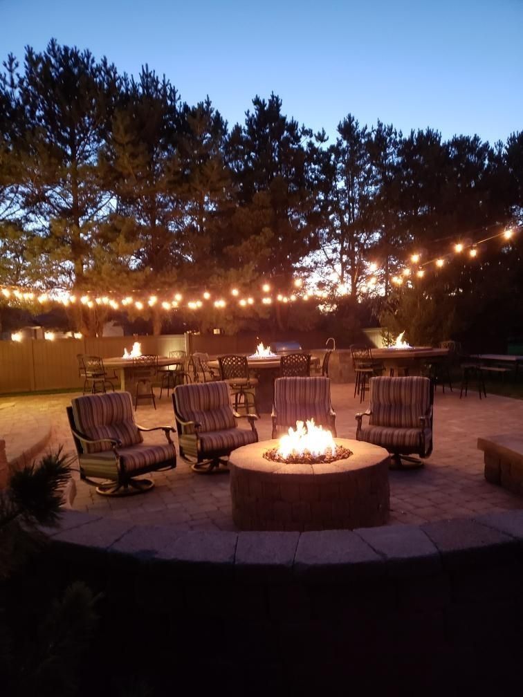 Outdoor patio at dusk with fire pit, seating, tables, string lights, and trees.