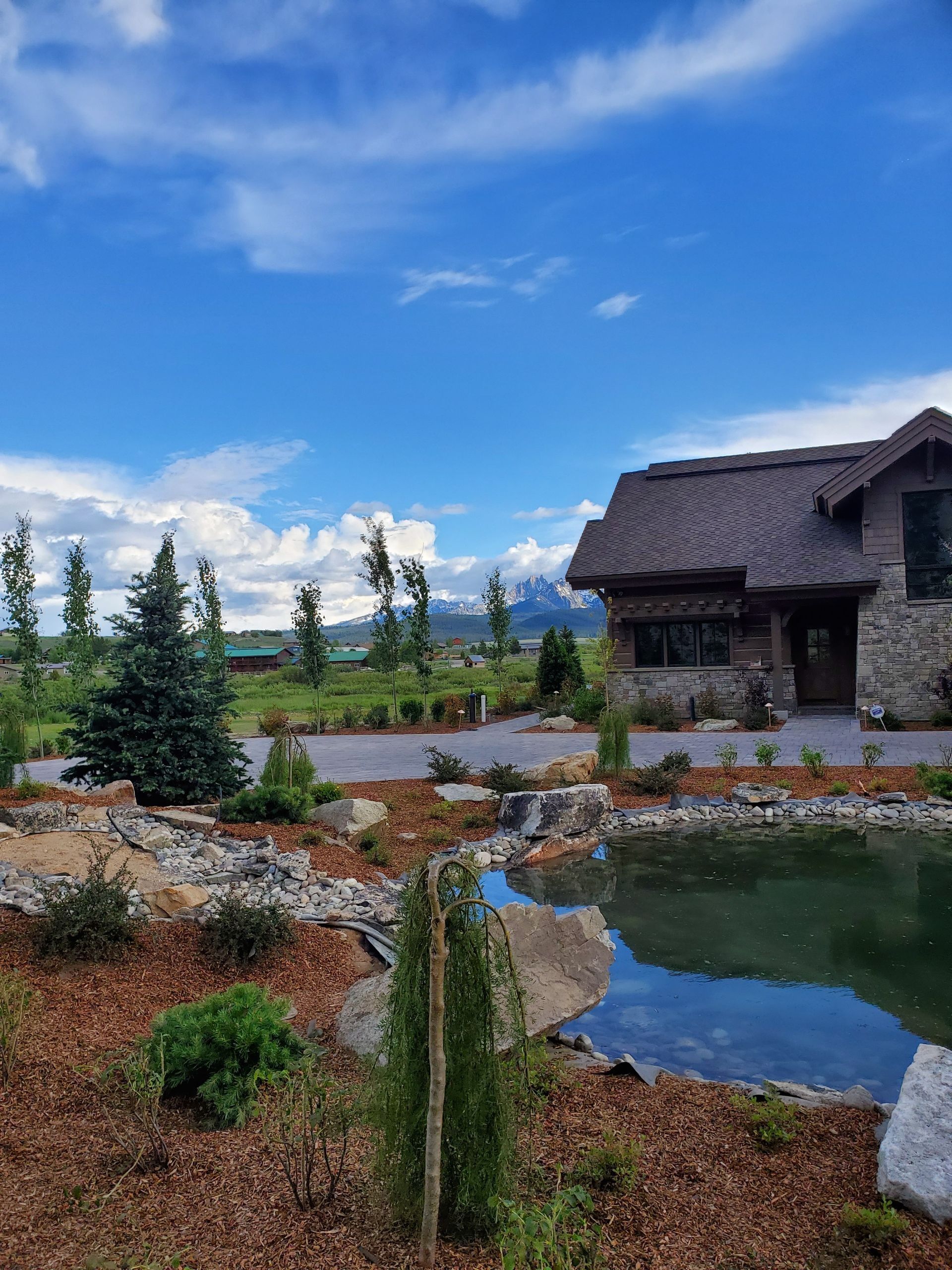 Stone house with thatched roof, pond, and mountains under a blue sky with clouds.