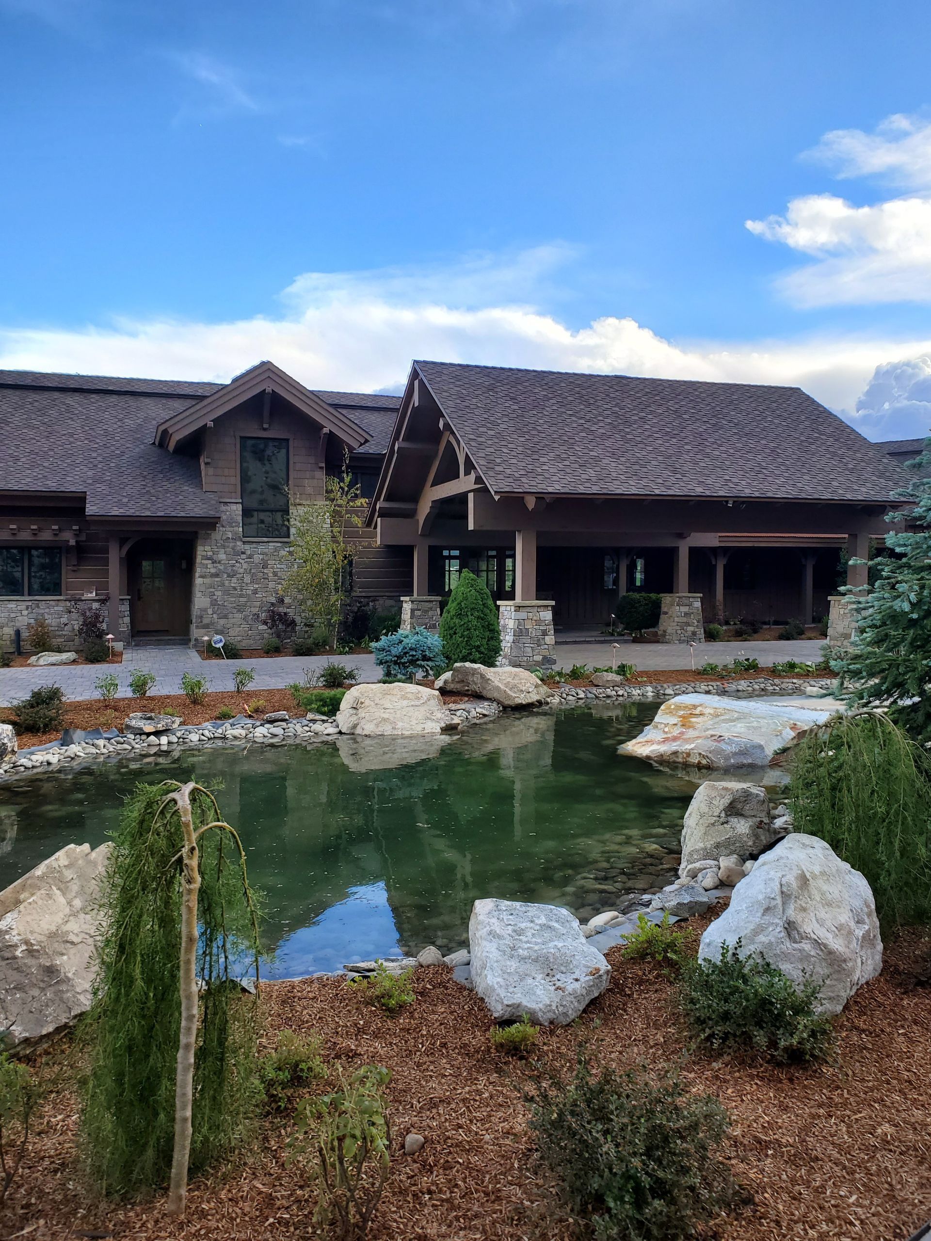 Lodge-style building with a pond in front. Brown shingle roof, stone pillars. Blue sky with clouds.