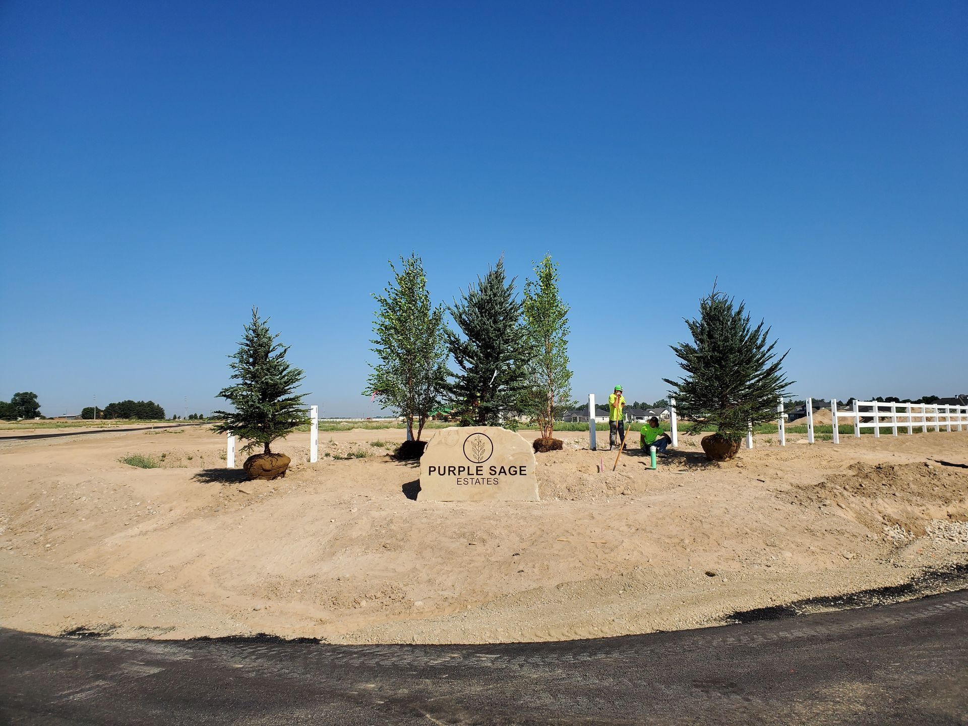 Sign with trees, landscaping in a rural area. Two people work, white fence and blue sky.