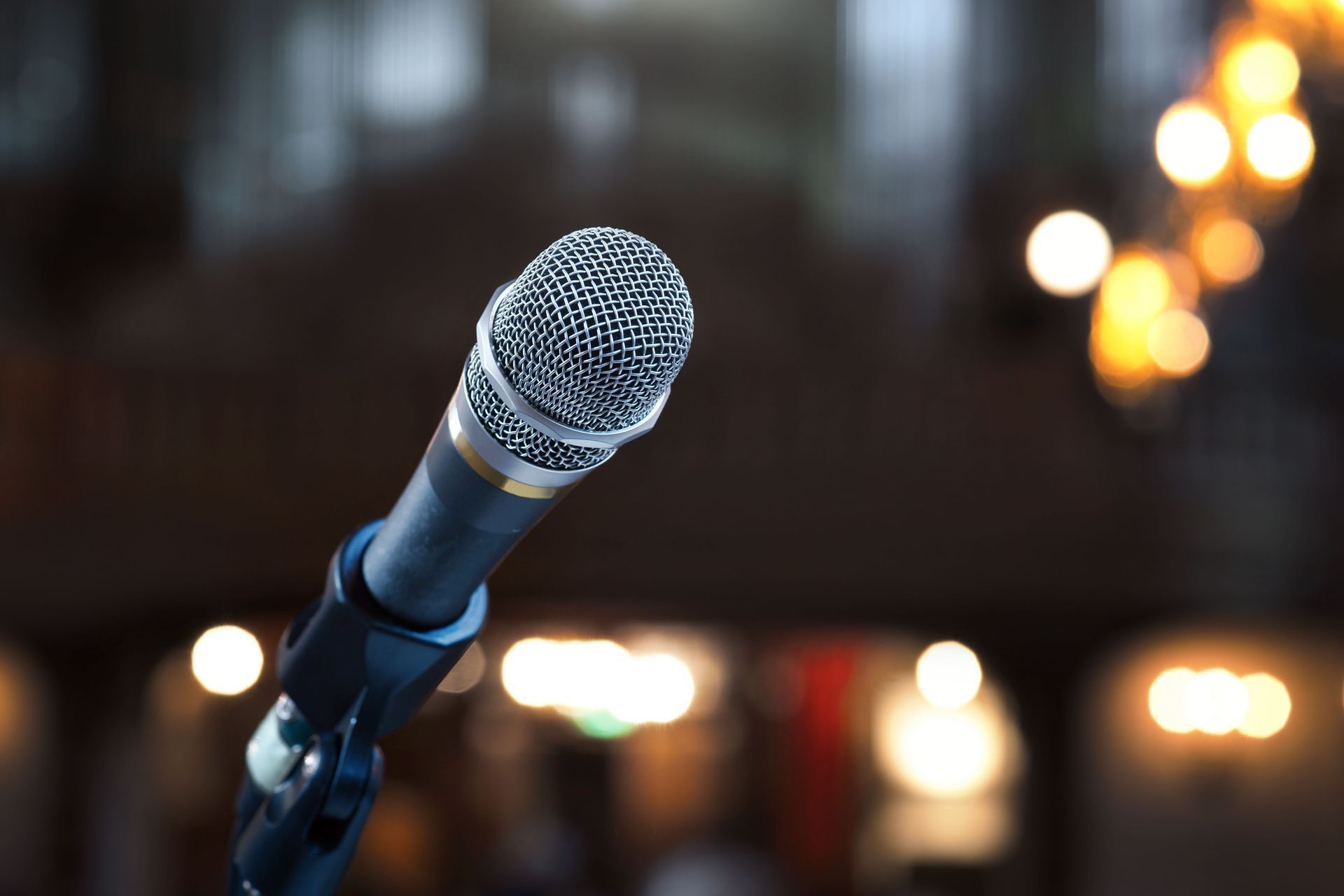 A close-up of a microphone on a stand against a blurred, dark background with warm, out-of-focus golden lights.