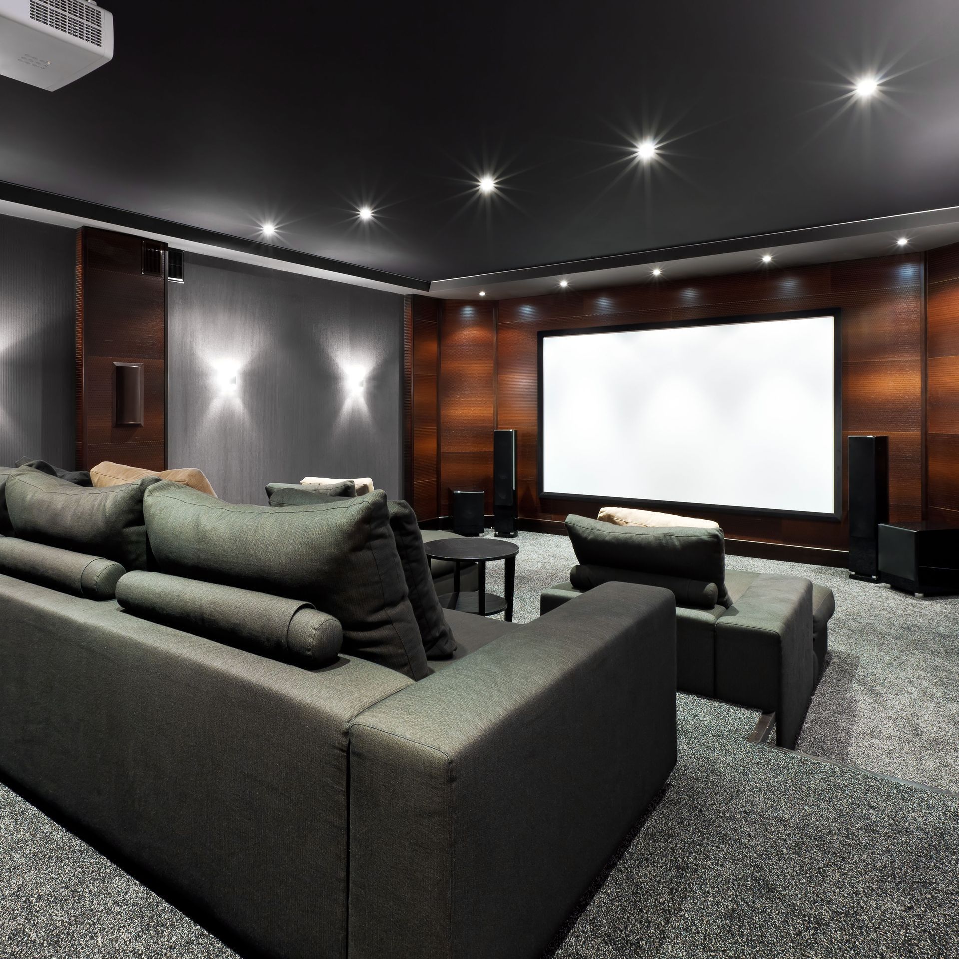 A modern home theater with gray sofas, wood-paneled walls, a large projection screen, and dim recessed lighting.
