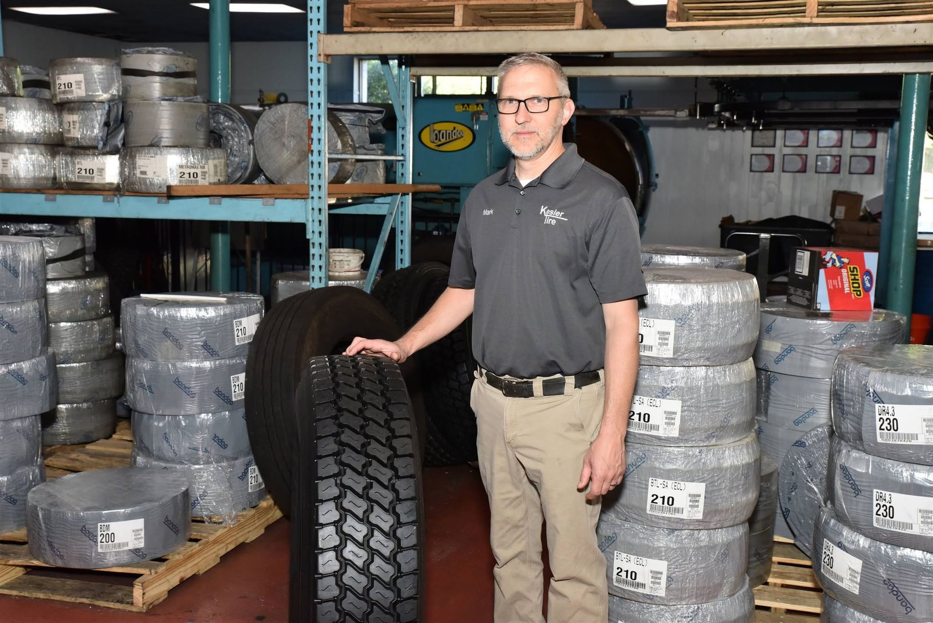 Man standing beside a large tire in a warehouse stacked with silver rims