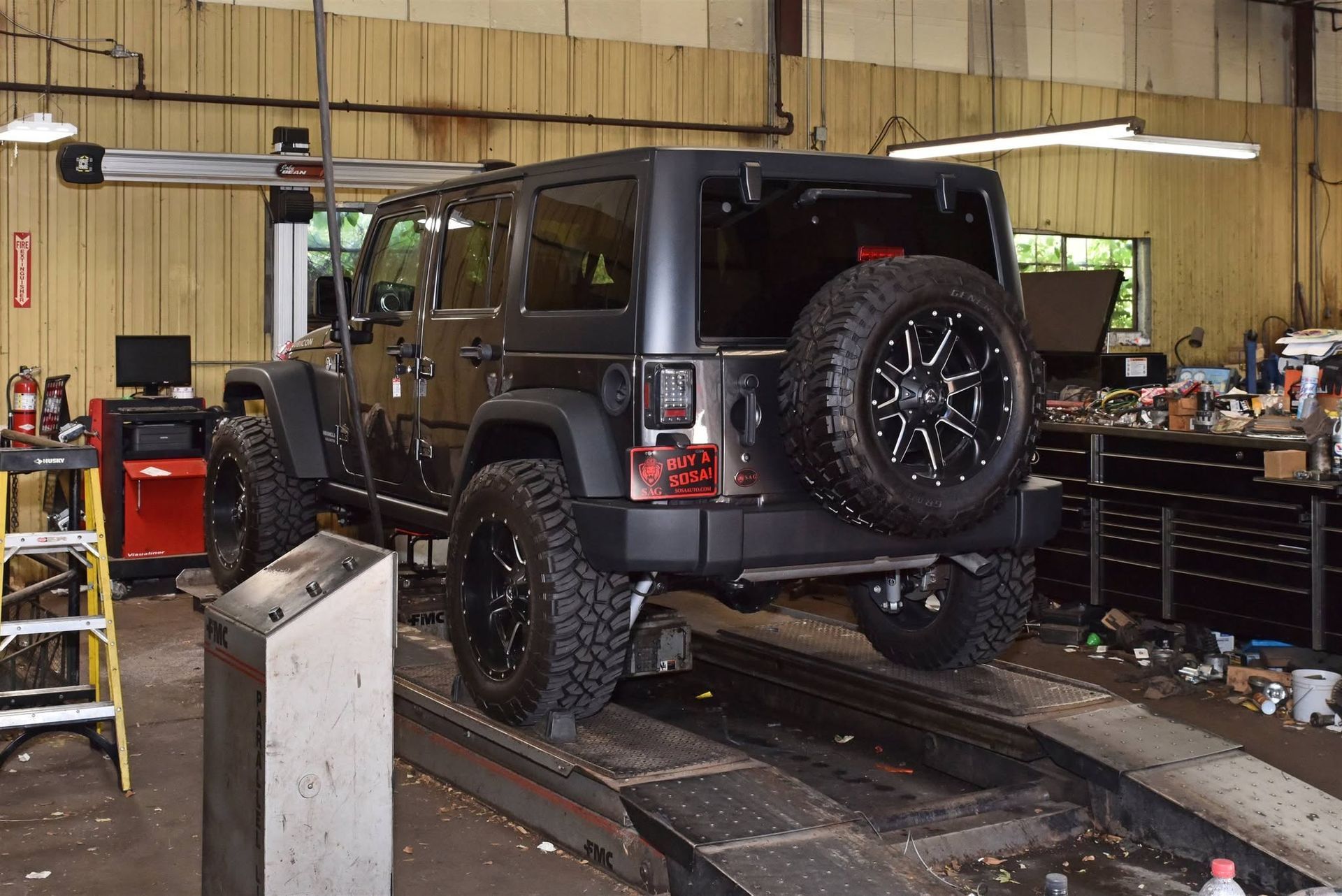 Black Jeep Wrangler on a lift inside an auto repair shop, viewed from the rear.