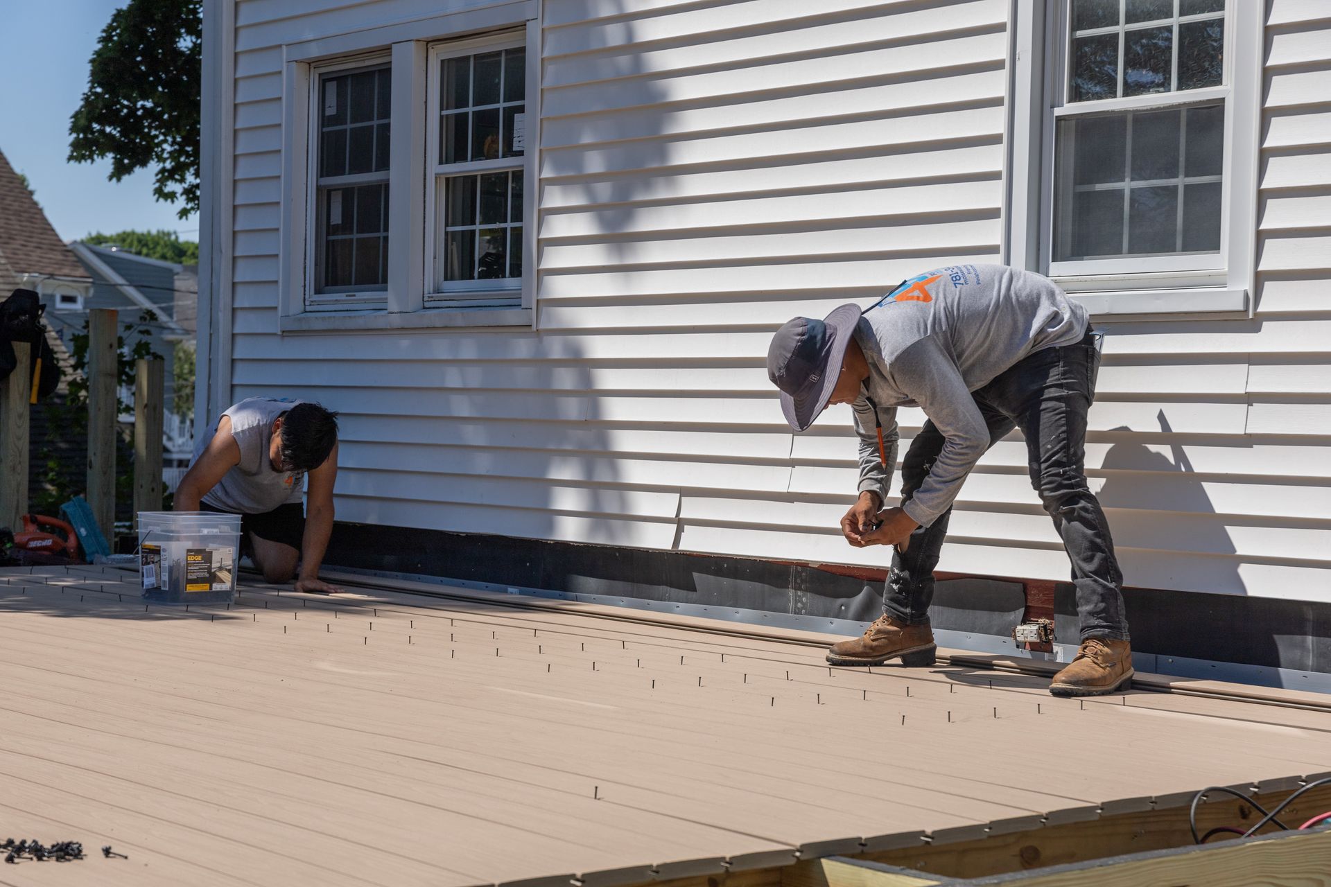 Two workers in hats and work clothes install light-colored composite decking planks outside a white-sided house.