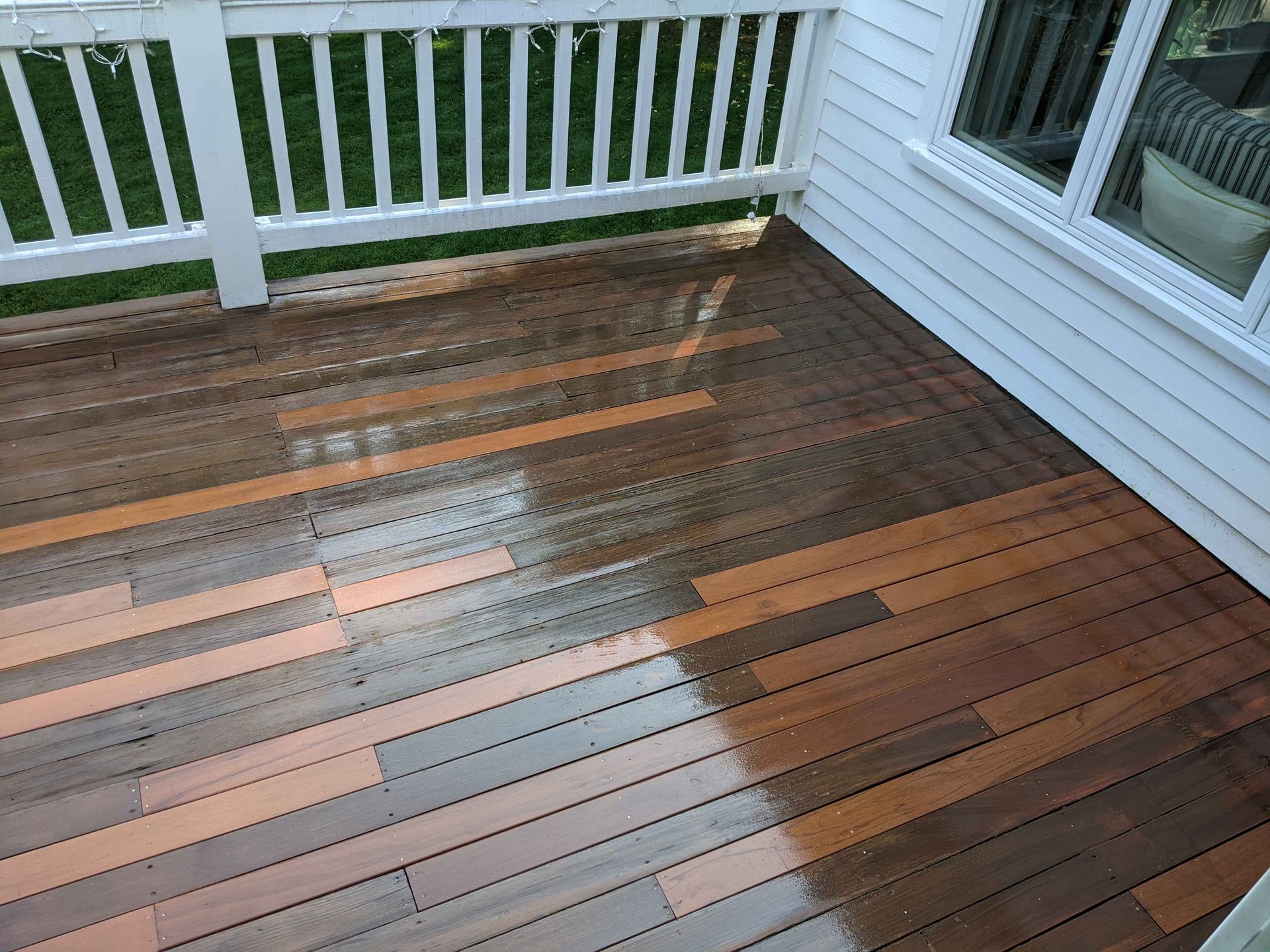 A wet wooden deck with a mix of light and dark brown boards, seen from a high angle next to a white house wall and railing.