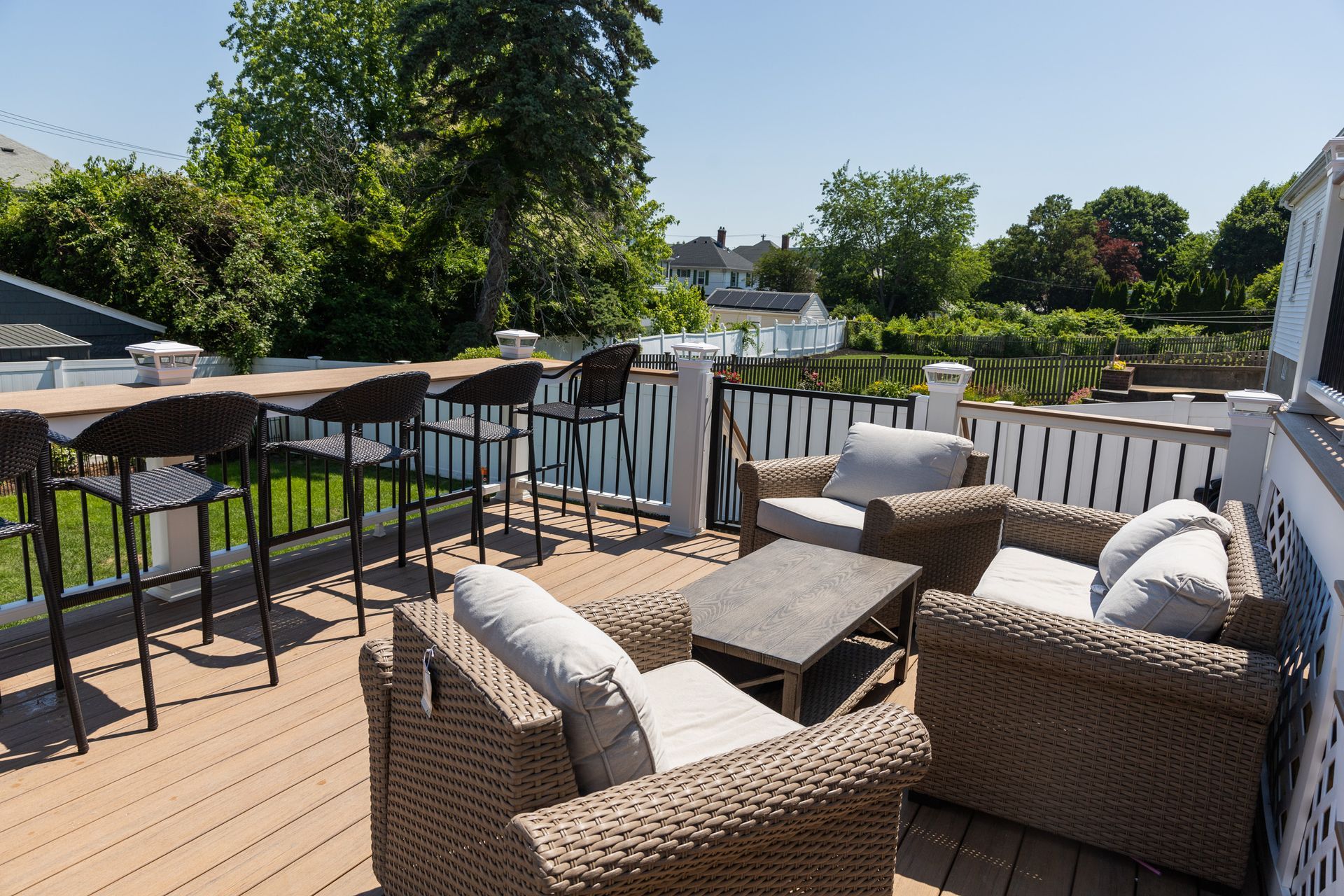 An outdoor wooden deck featuring wicker chairs, a coffee table, and a bar table with stools overlooking a sunny yard.
