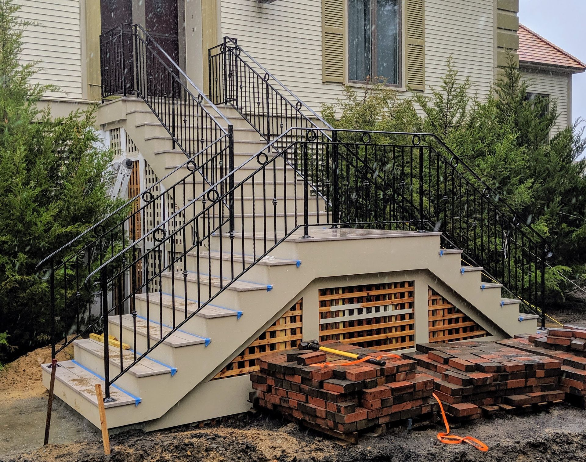 Outdoor concrete stairs with black iron railings leading up to a house entrance, surrounded by greenery and brick debris.