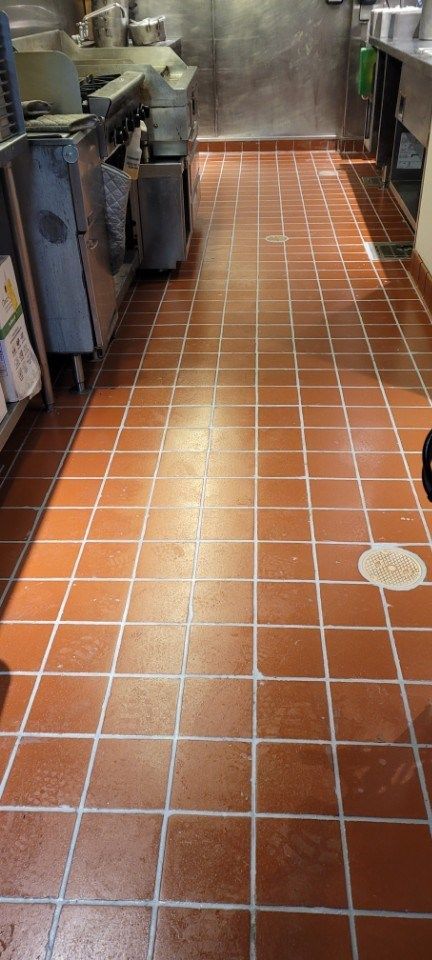 A narrow, commercial kitchen floor covered in red square tiles, with metal equipment lining the left wall.