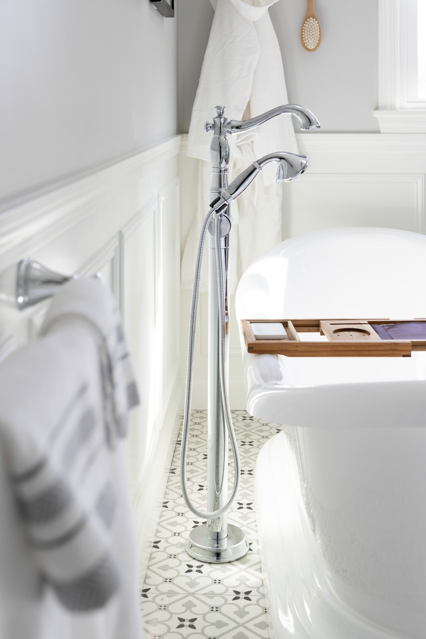 A chrome floor-mounted bathtub faucet with a handheld shower sprayer beside a white soaking tub in a bright bathroom.