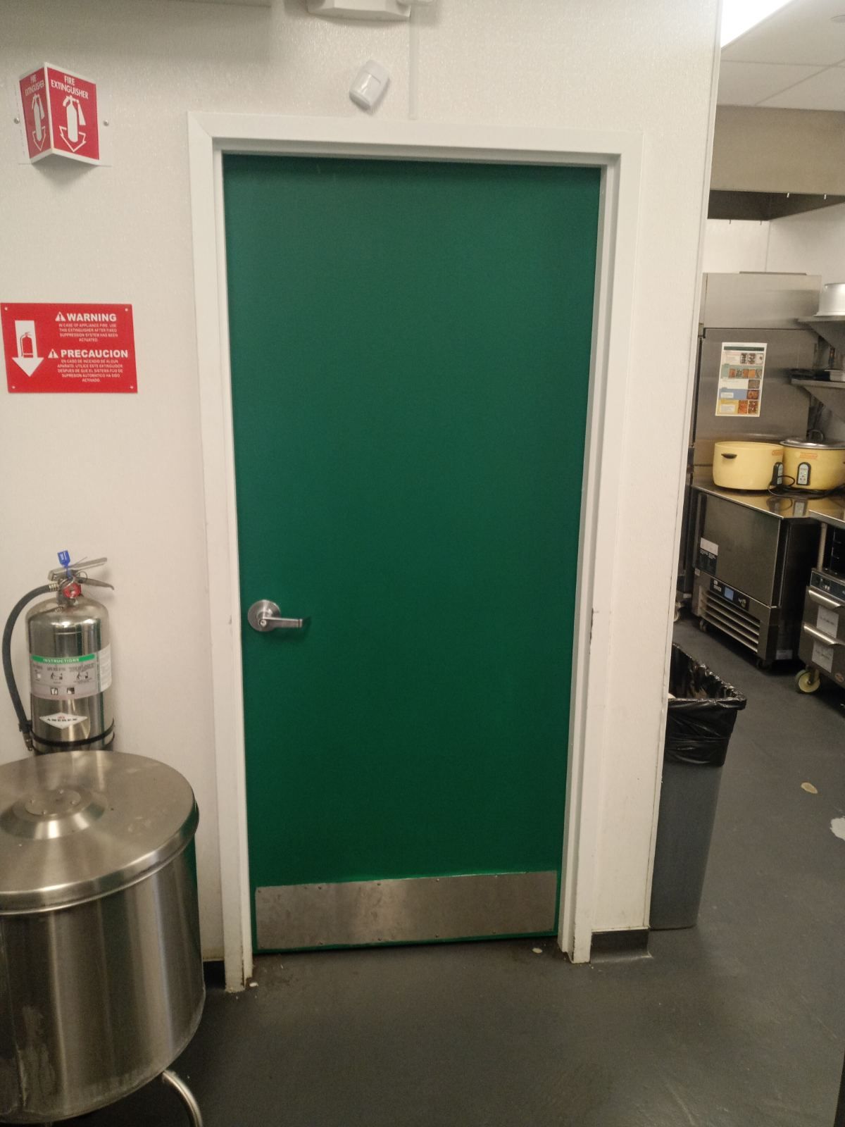 A green door with a metal kick plate in a commercial kitchen next to a fire extinguisher and signage on a white wall.