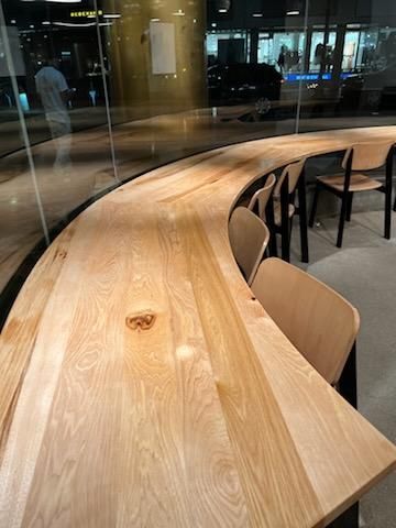 A curved, light-wood counter sits next to a glass window in a modern cafe, accompanied by matching wooden chairs.