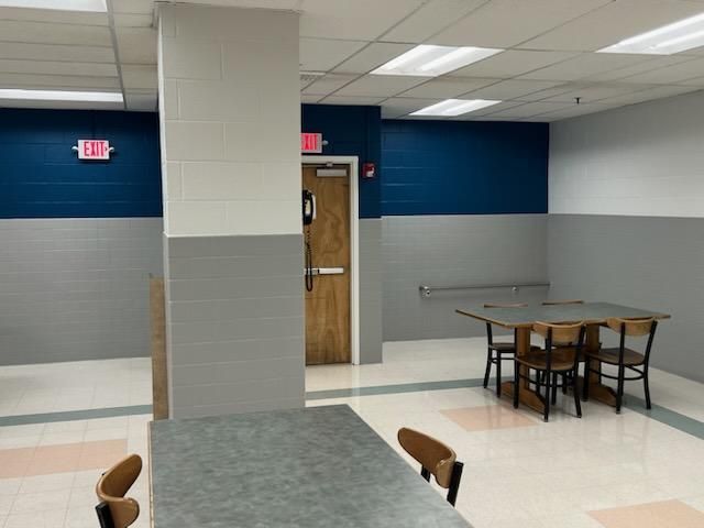 A cafeteria space featuring tables and chairs, with gray and navy blue walls and a wooden door near a central pillar.