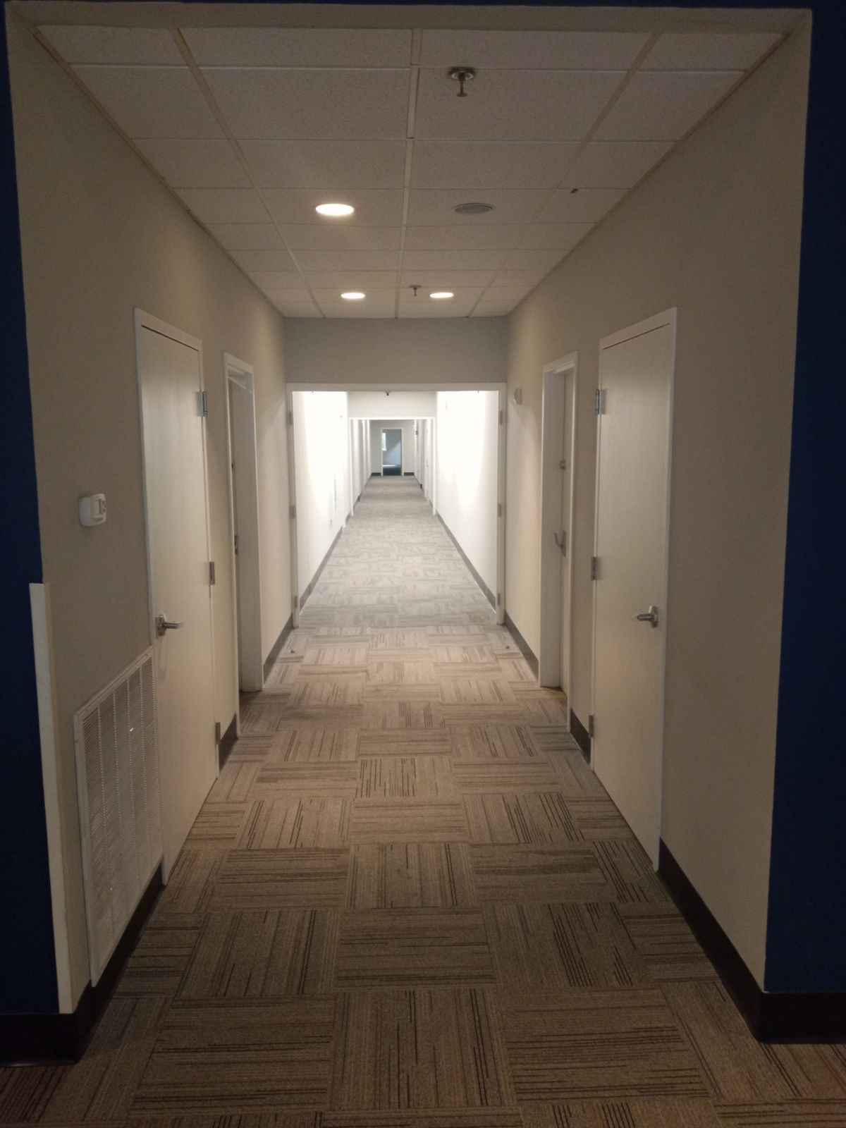 A long, straight office hallway with neutral walls, light-colored carpet tiles, and multiple closed white doors.
